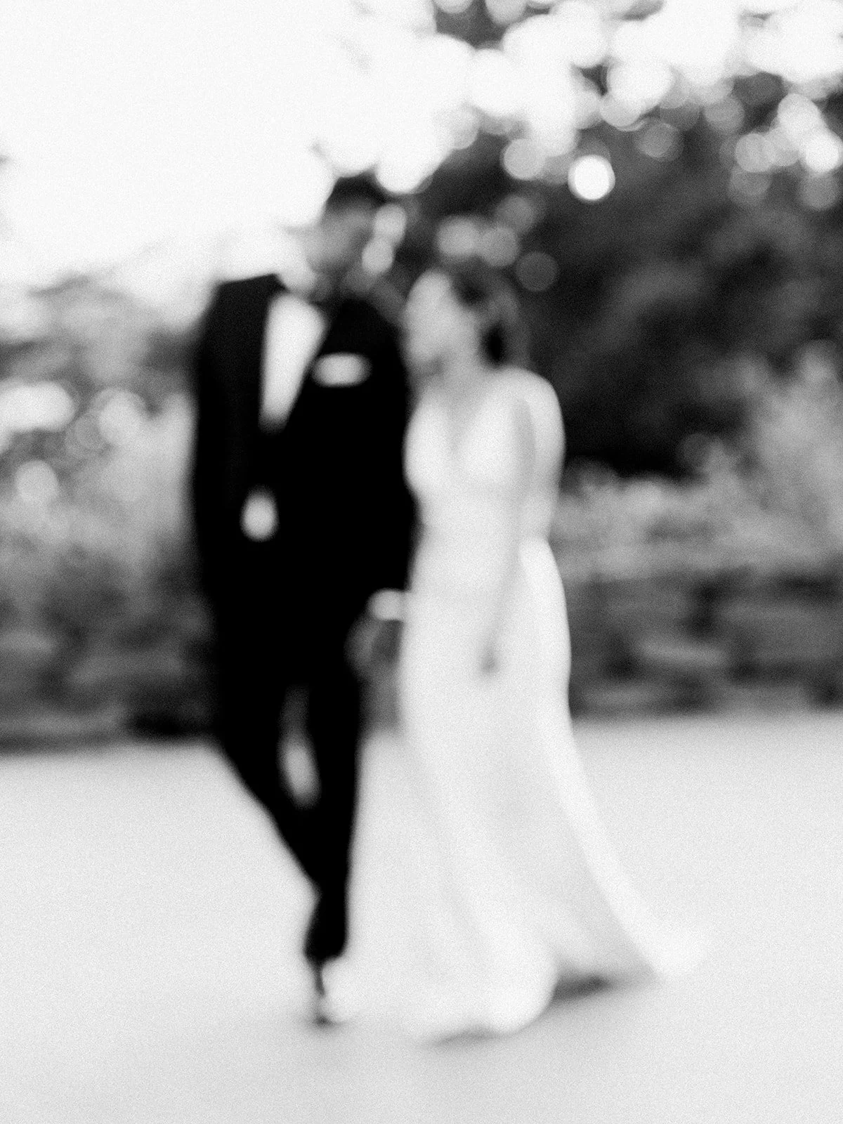 Blurry black and white photo of a bride and groom standing outdoors, dressed in wedding attire, with trees or bushes in the background.