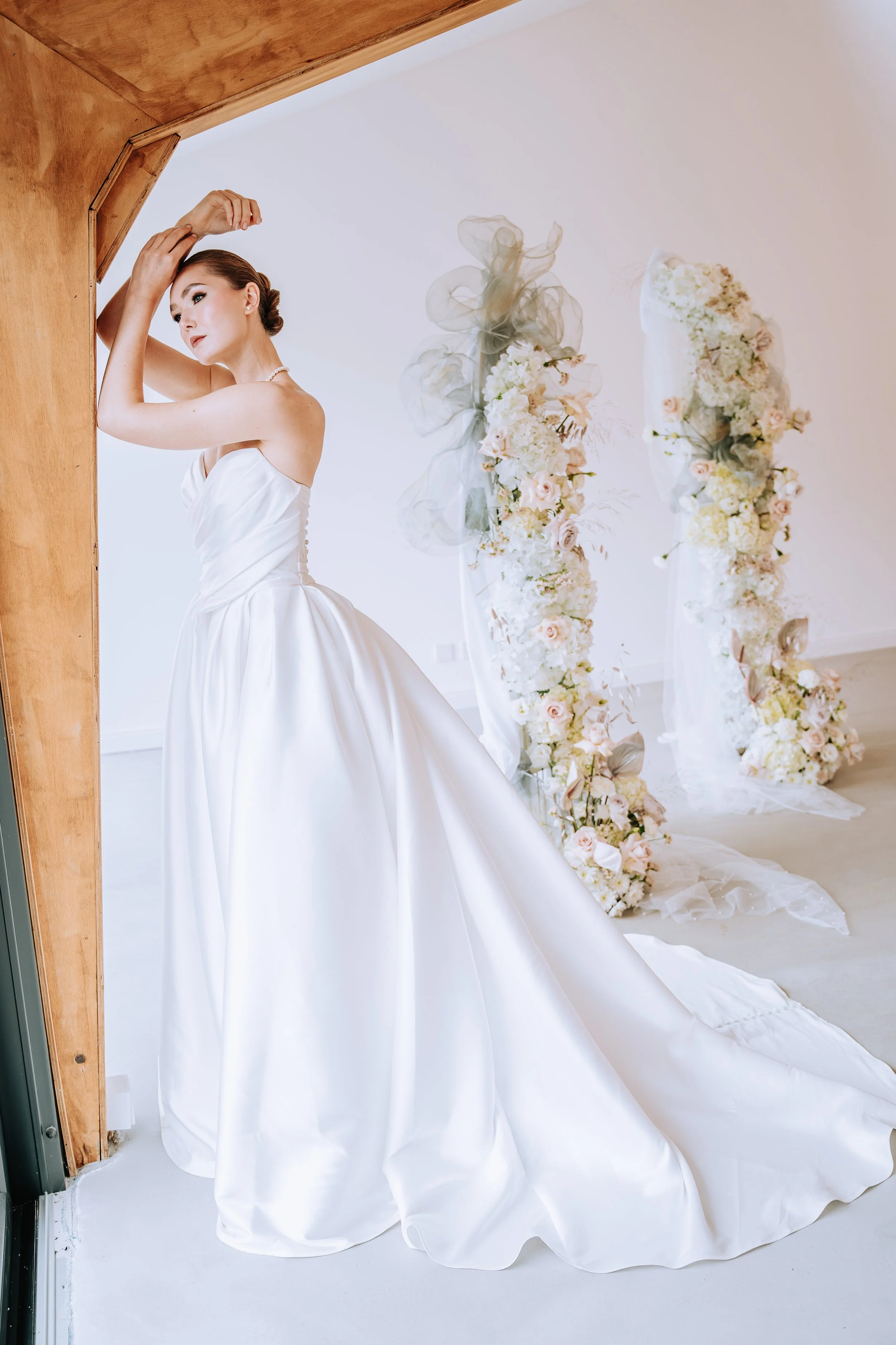 Braut in weißem Hochzeitskleid steht in einem Raum mit Blumen und Dekorationen im Hintergrund.
