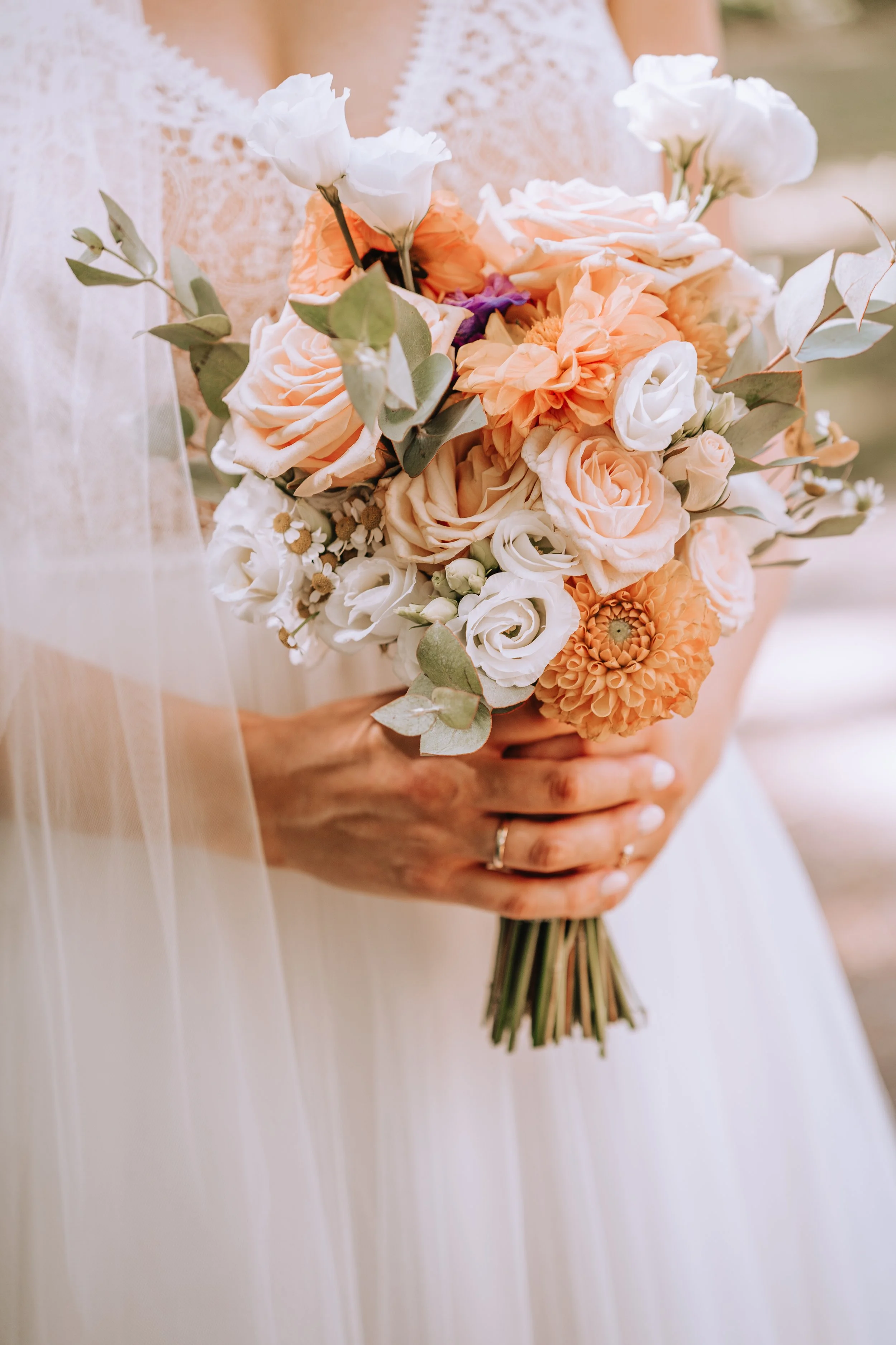 Eine Braut hält einen Blumenstrauß mit weißen, peachfarbenen und orangenen Blumen, umgeben von grünen Blättern.