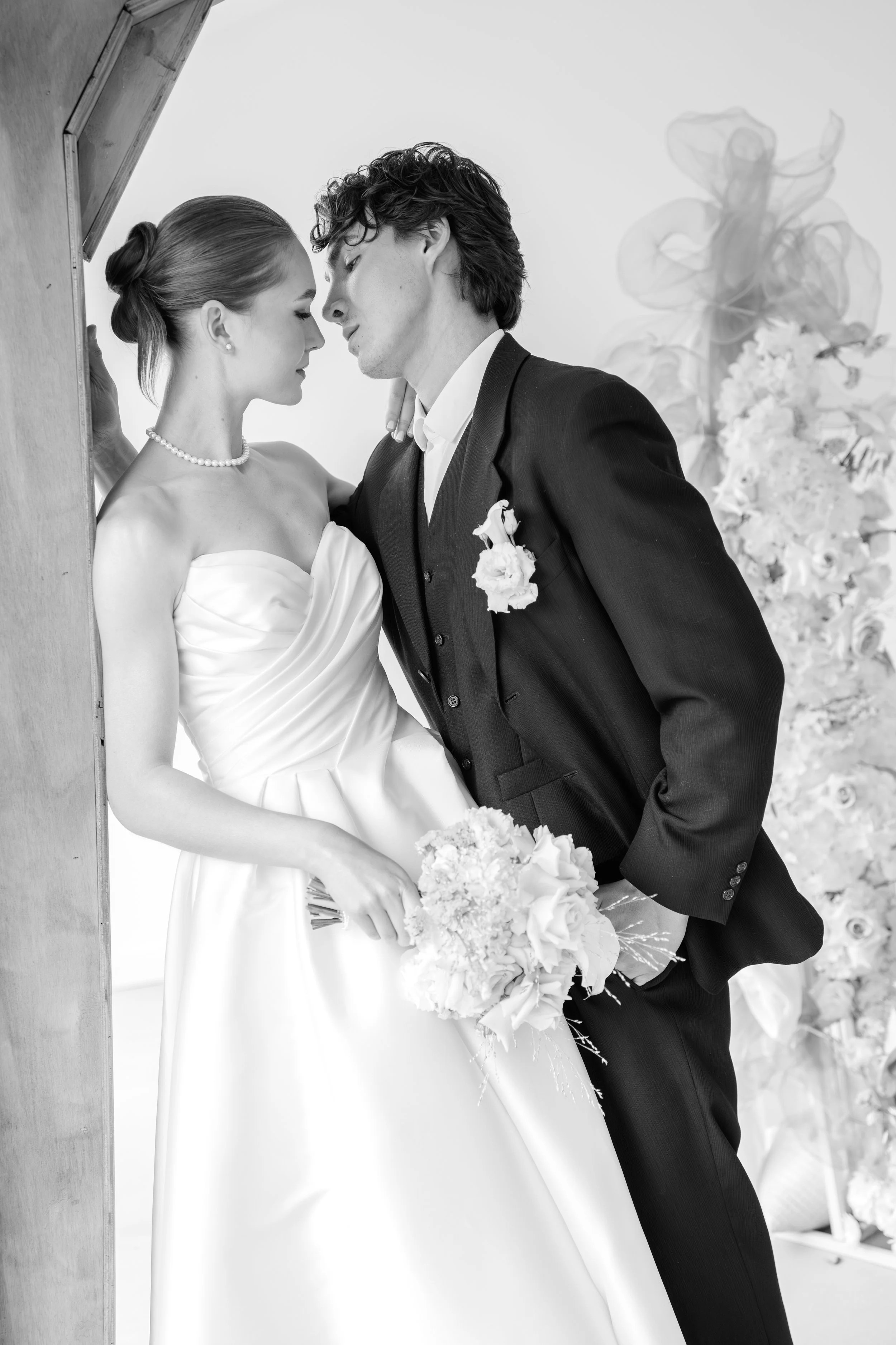 Braut und Bräutigam beim Hochzeitsfotoshooting, stehen nahe beieinander, mit Blumen vor einem Hintergrund aus Blumen und Dekorationen, in Schwarz-Weiß aufgenommen.