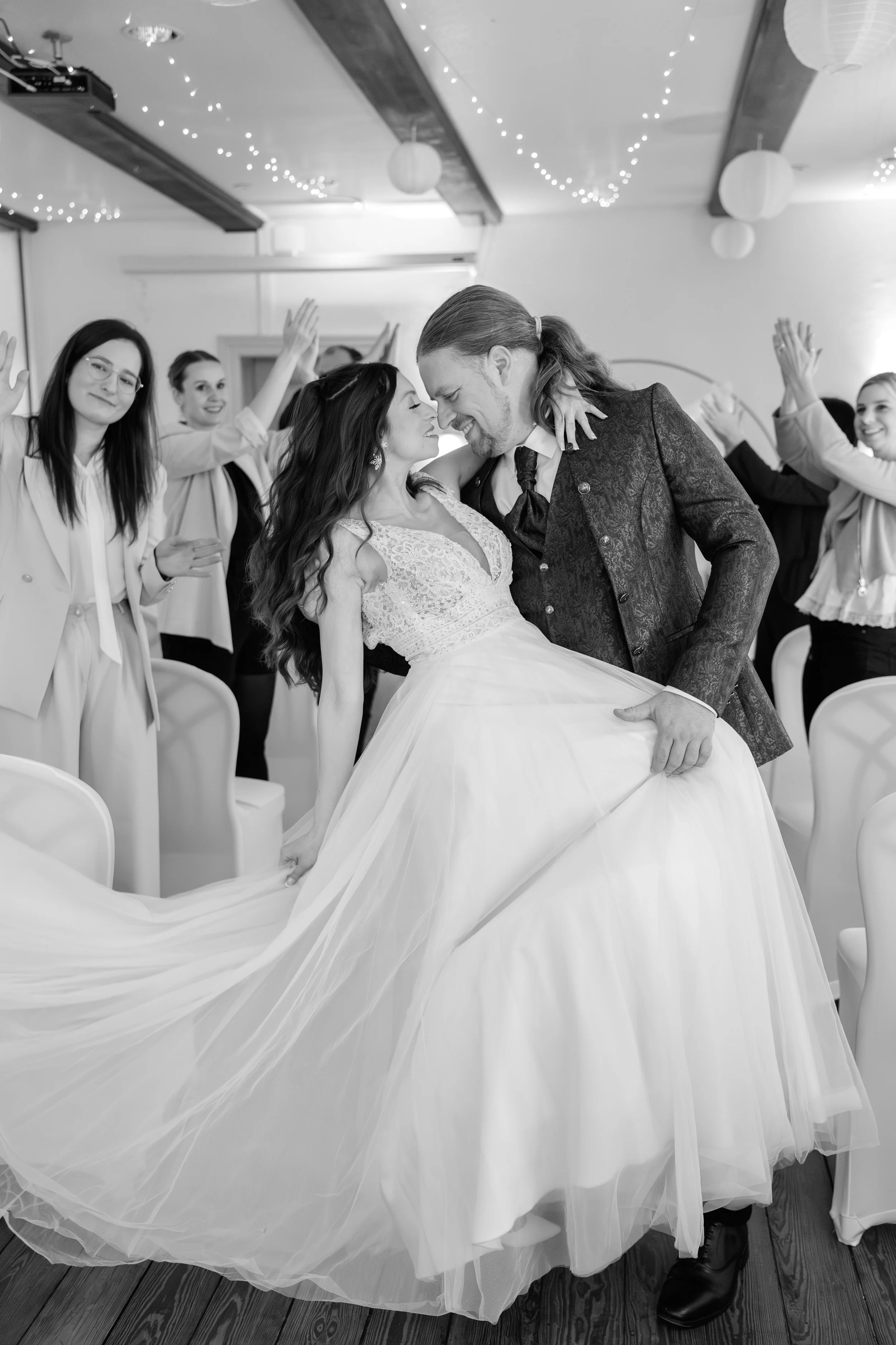 Ein Brautpaar tanzt bei einer Hochzeit, umgeben von lachenden Freunden, in einer festlich dekorierten Halle mit Lichterketten und Papierlampions.