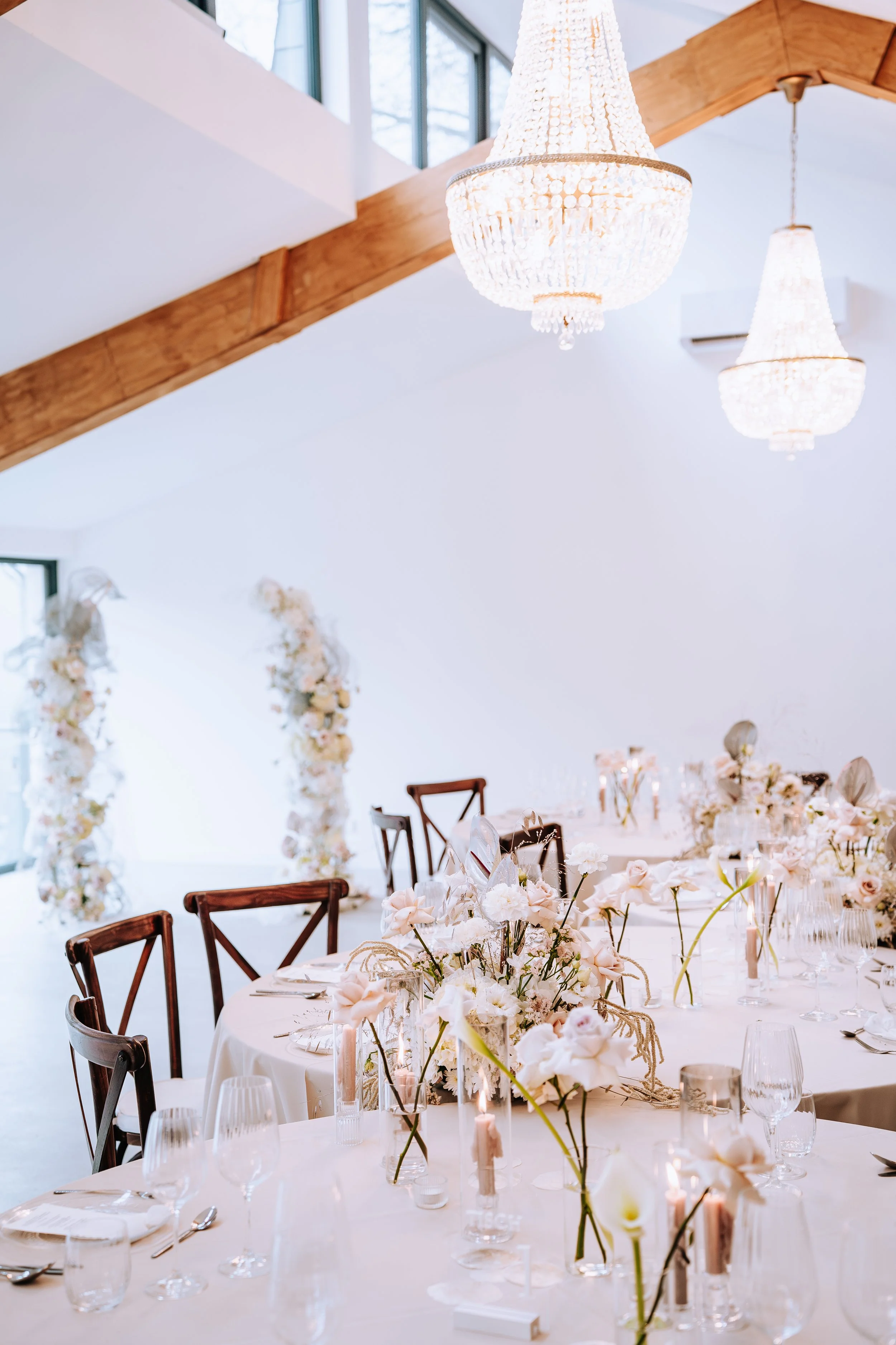 Eleganter festlicher Raum mit runden Tischen, gedeckt mit weißen Tuchhussen, geschmückt mit Blumen, Kerzen und Gläsern, Chandeliers an der Decke und große Fenster mit natürlichem Licht.