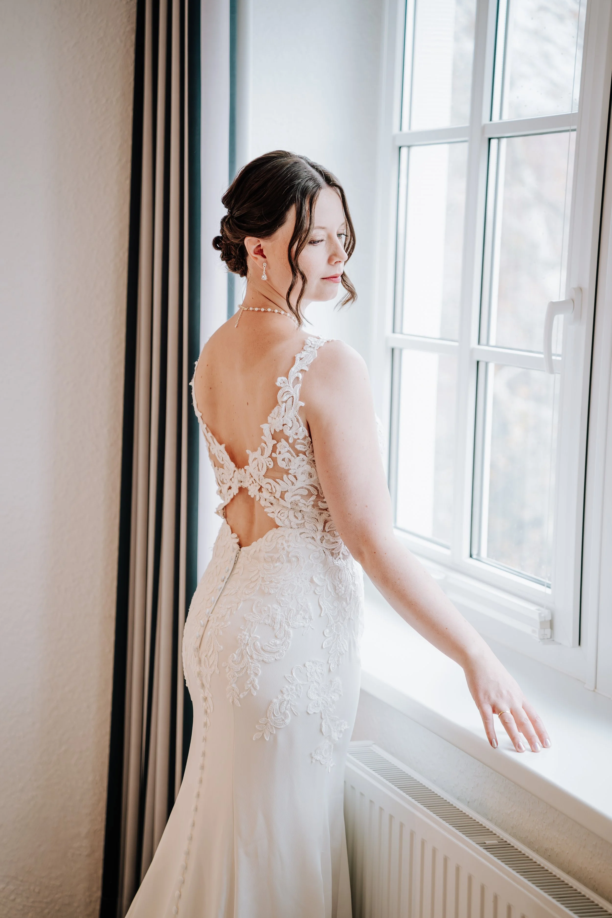 Braut in weißem Hochzeitskleid schaut aus dem Fenster.