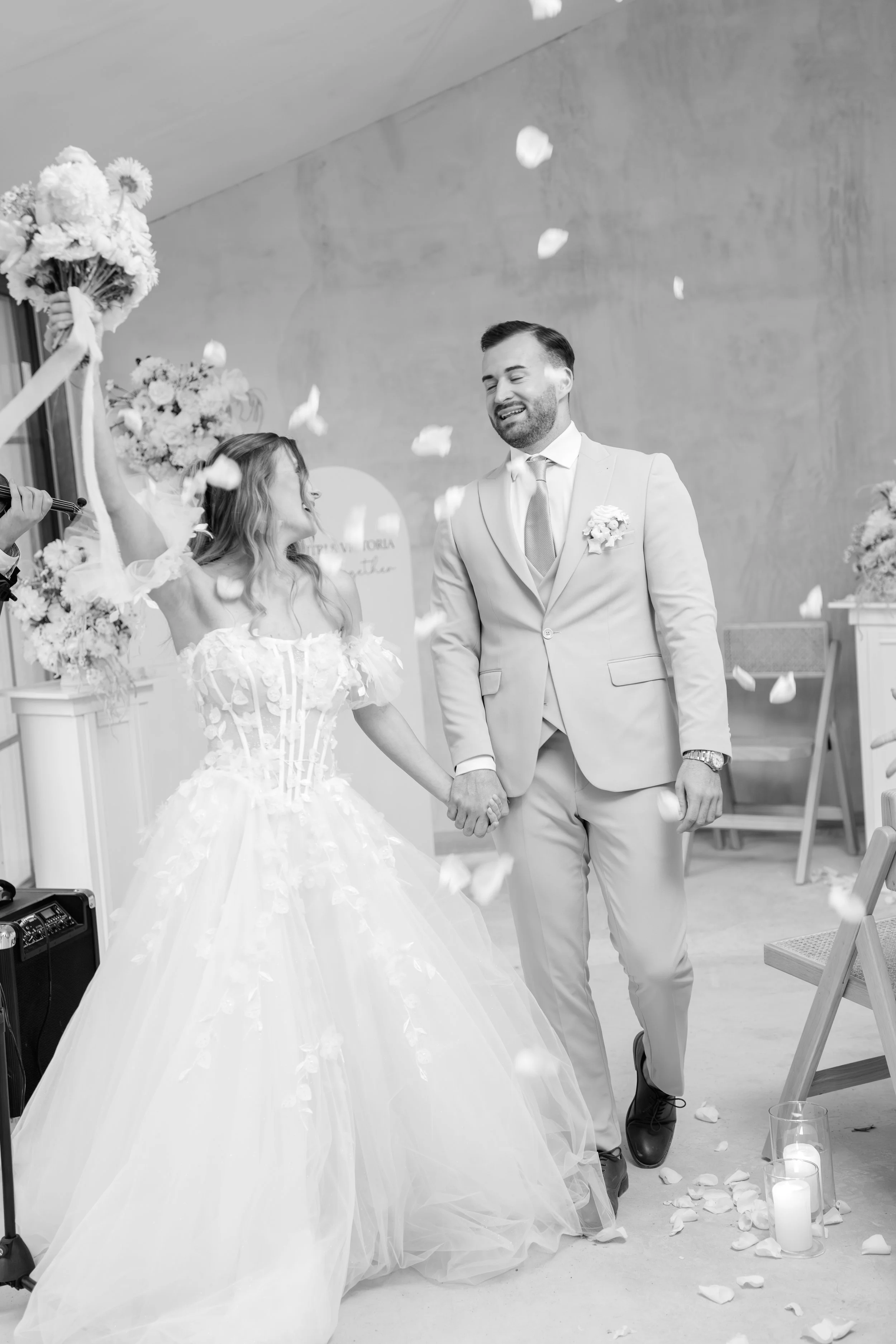 Braut und Bräutigam bei ihrer Hochzeit, Hand in Hand, lächelnd, mit Blumen und Kerzen im Hintergrund, in Schwarz-Weiß aufgenommen.