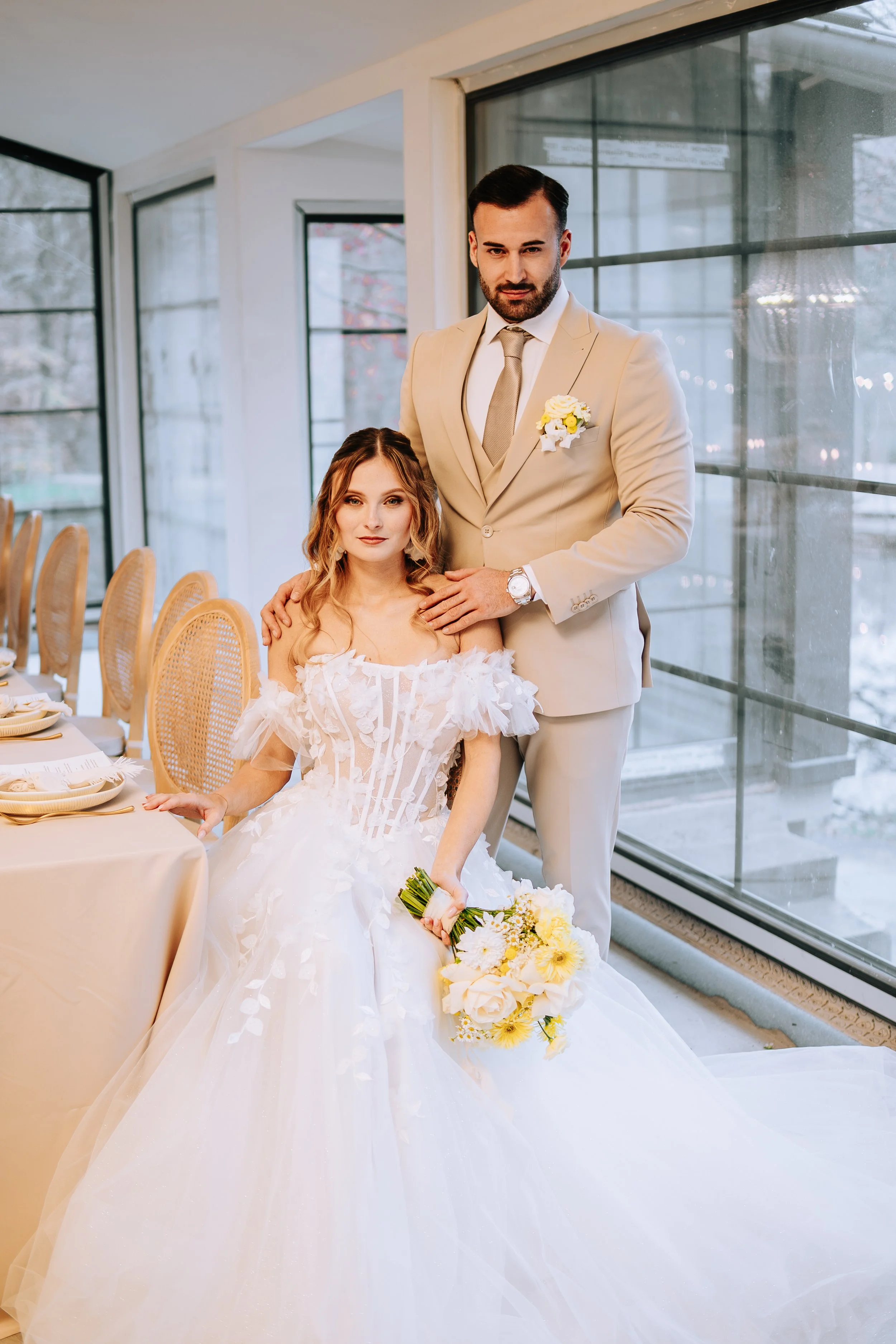 Ein Brautpaar bei einer Hochzeit in einem hell erleuchteten Raum, der Bräutigam trägt einen beigen Anzug mit Krawatte und Blumenboutonnière, die Braut trägt ein weißes Hochzeitskleid mit floralen Details und hält einen Blumenstrauß, sie sitzen an ein