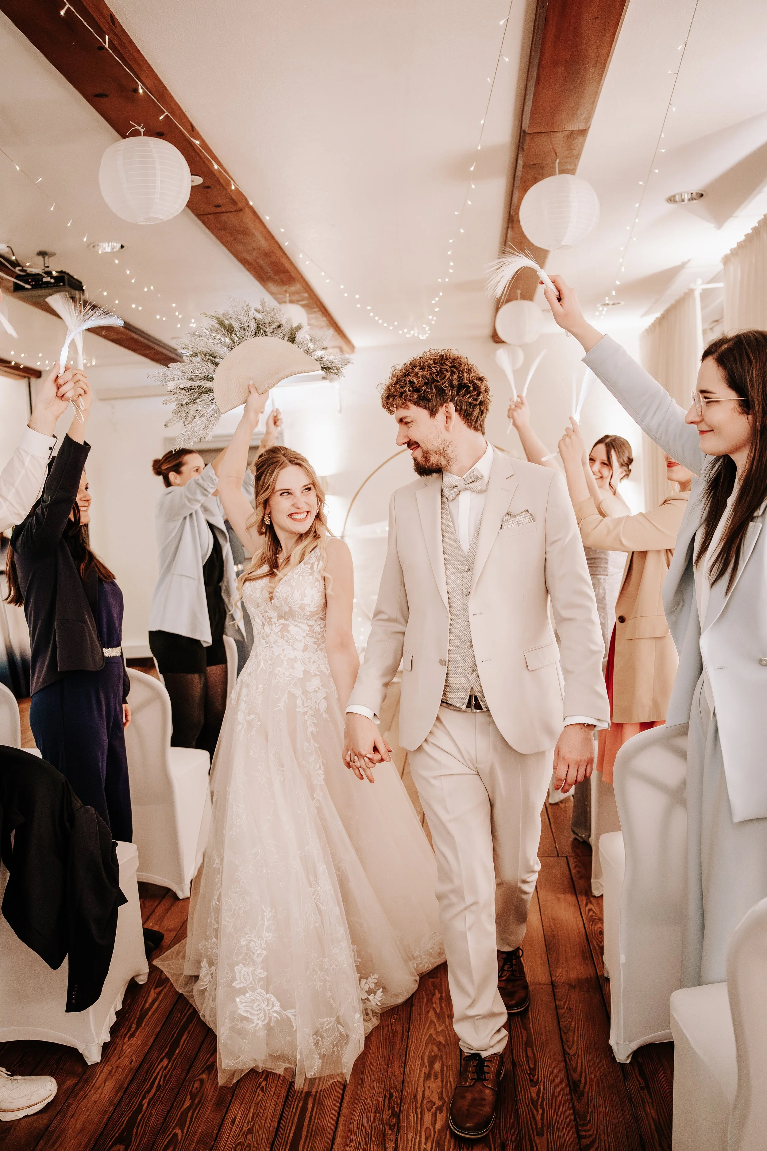 Ein Brautpaar läuft Hand in Hand durch eine festlich geschmückte Hochzeitsgesellschaft, die mit Papierlaternen und Lichterketten dekoriert ist, während die Gäste feierlich mit Federschlägern und Hüten applaudieren.