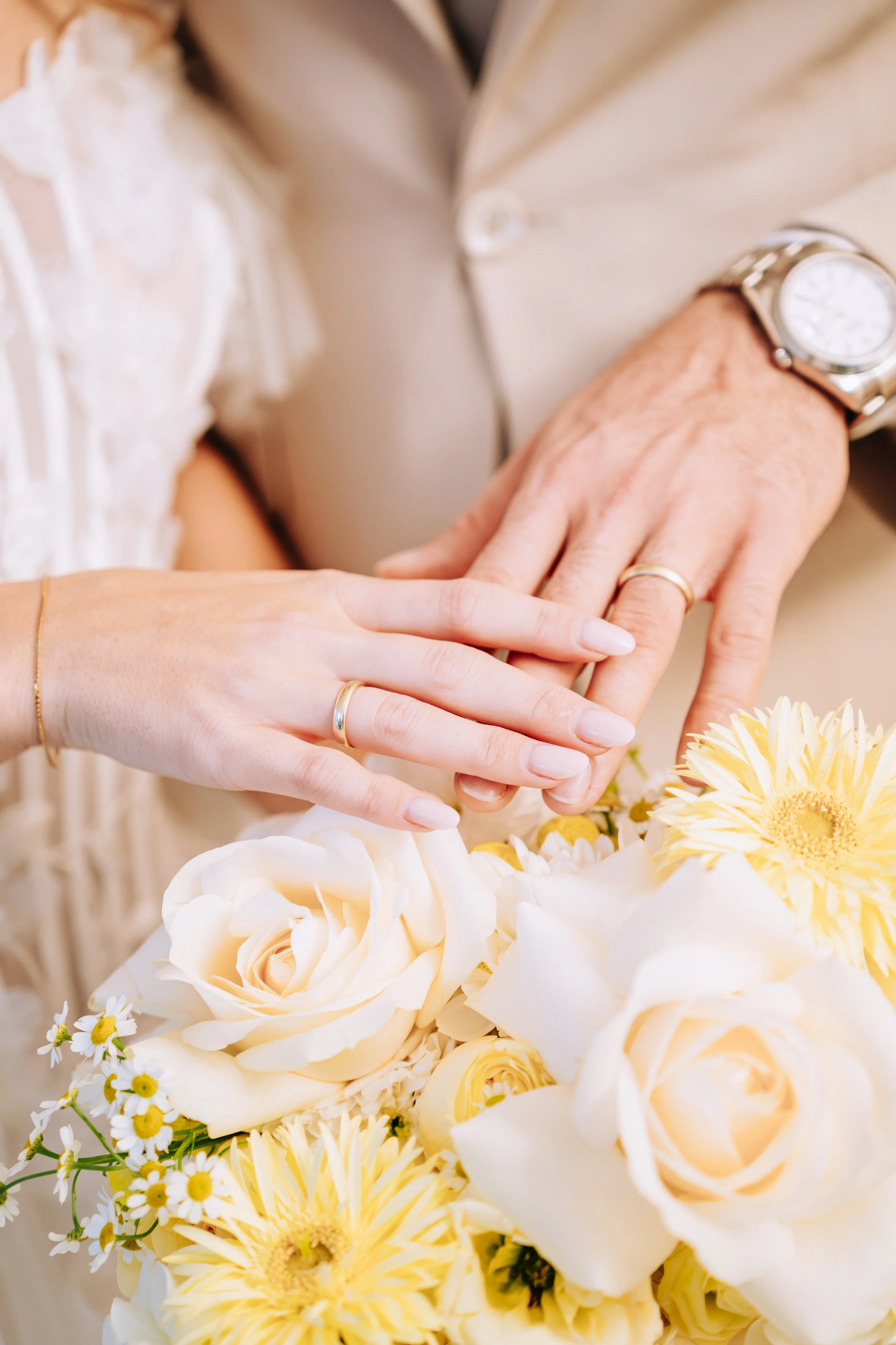 Nahaufnahme der Hände eines Paares, die auf einem Blumenstrauß liegen, mit Eheringen, im Hintergrund ist ein Teil eines Brautkleides und Anzugs sichtbar.