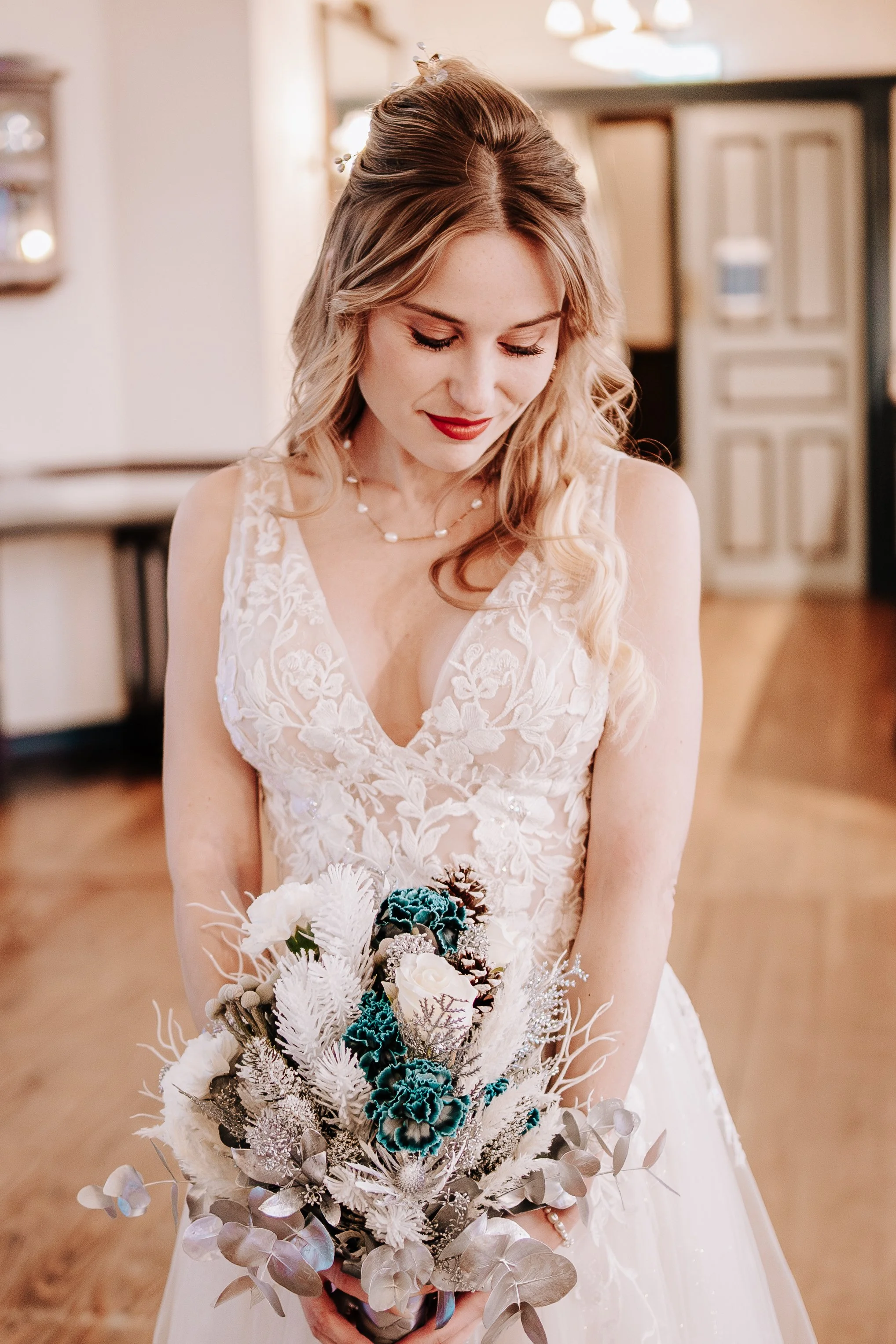 Braut in Hochzeitskleid hält einen Blumenstrauß, lächelt und blickt nach unten, im Hintergrund ein Raum mit Holzboden und Möbeln.
