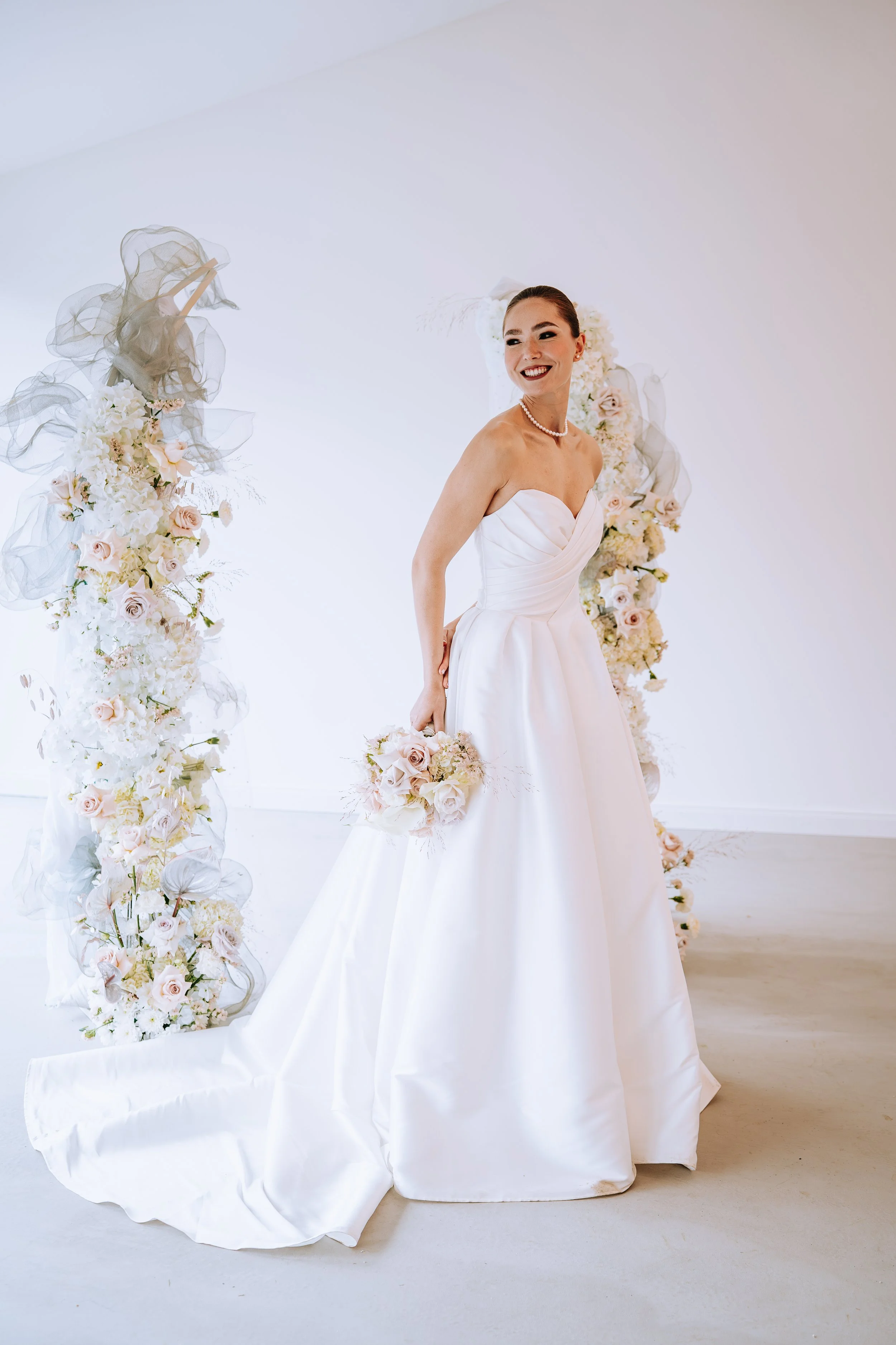 Braut in weißem Hochzeitskleid mit Blumenstrauß vor floral geschmückter Kulisse.