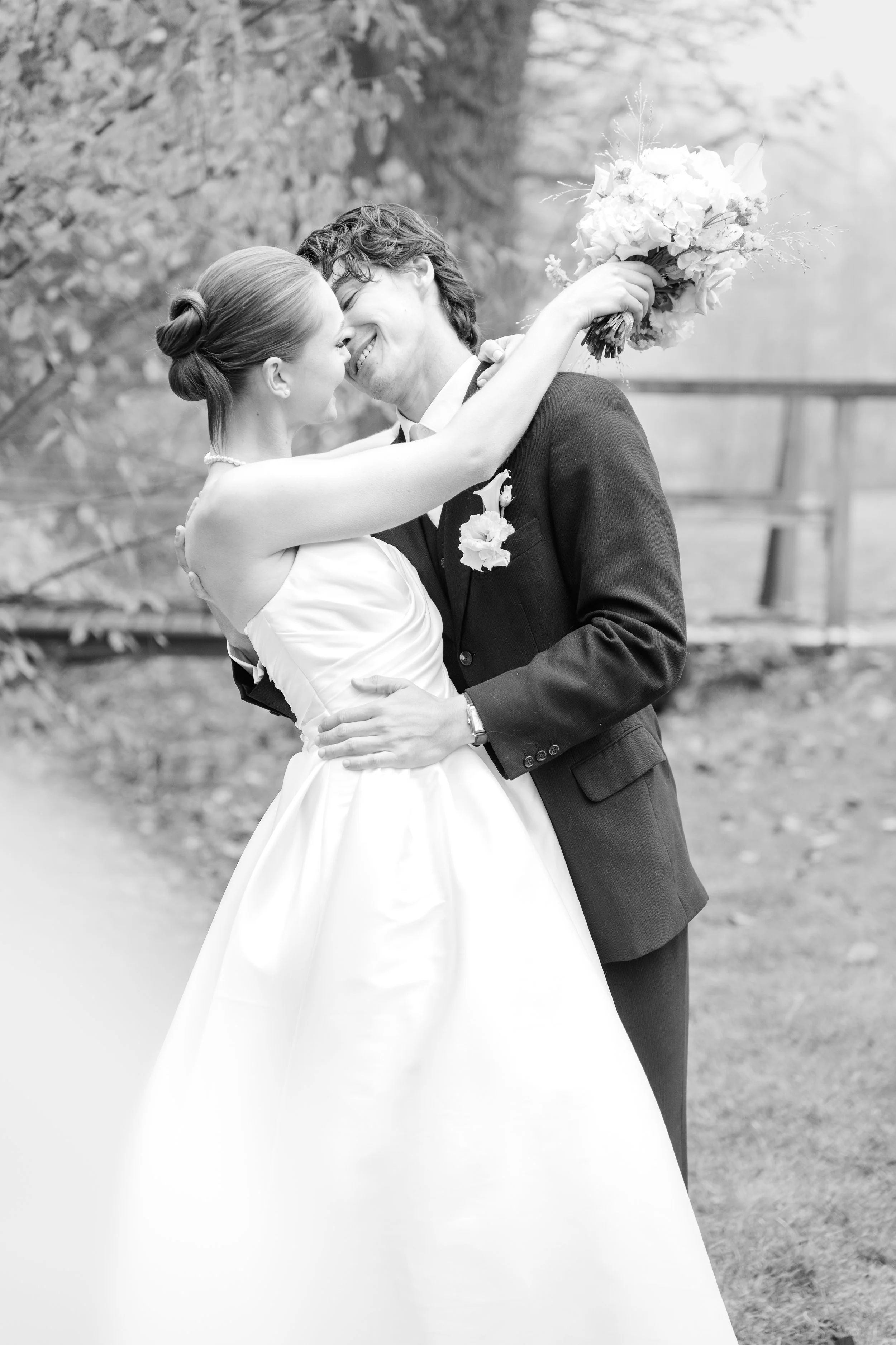 Ein Hochzeitspaar tanzt im Freien, sie umarmt ihn und hält einen Blumenstrauß in der Hand, beide lächeln sich glücklich an.