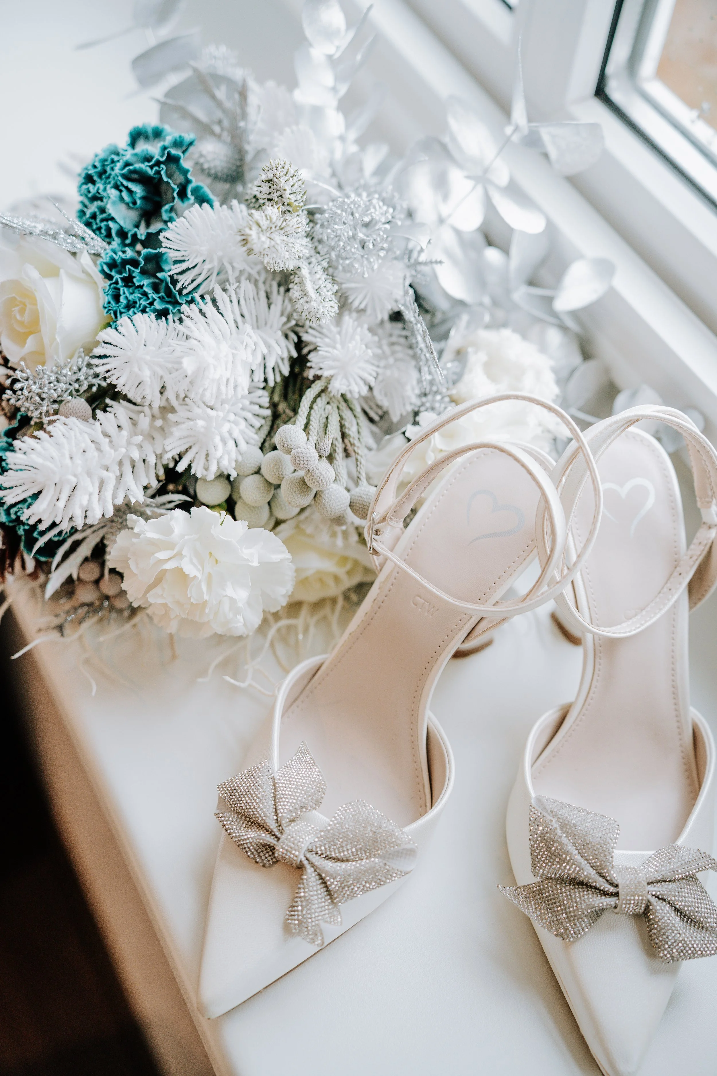 Weiße High-Heels mit silbernen Schleifen neben einem Blumenstrauß mit weißen und blauen Dekorationen, auf einer weißen Fensterbank.