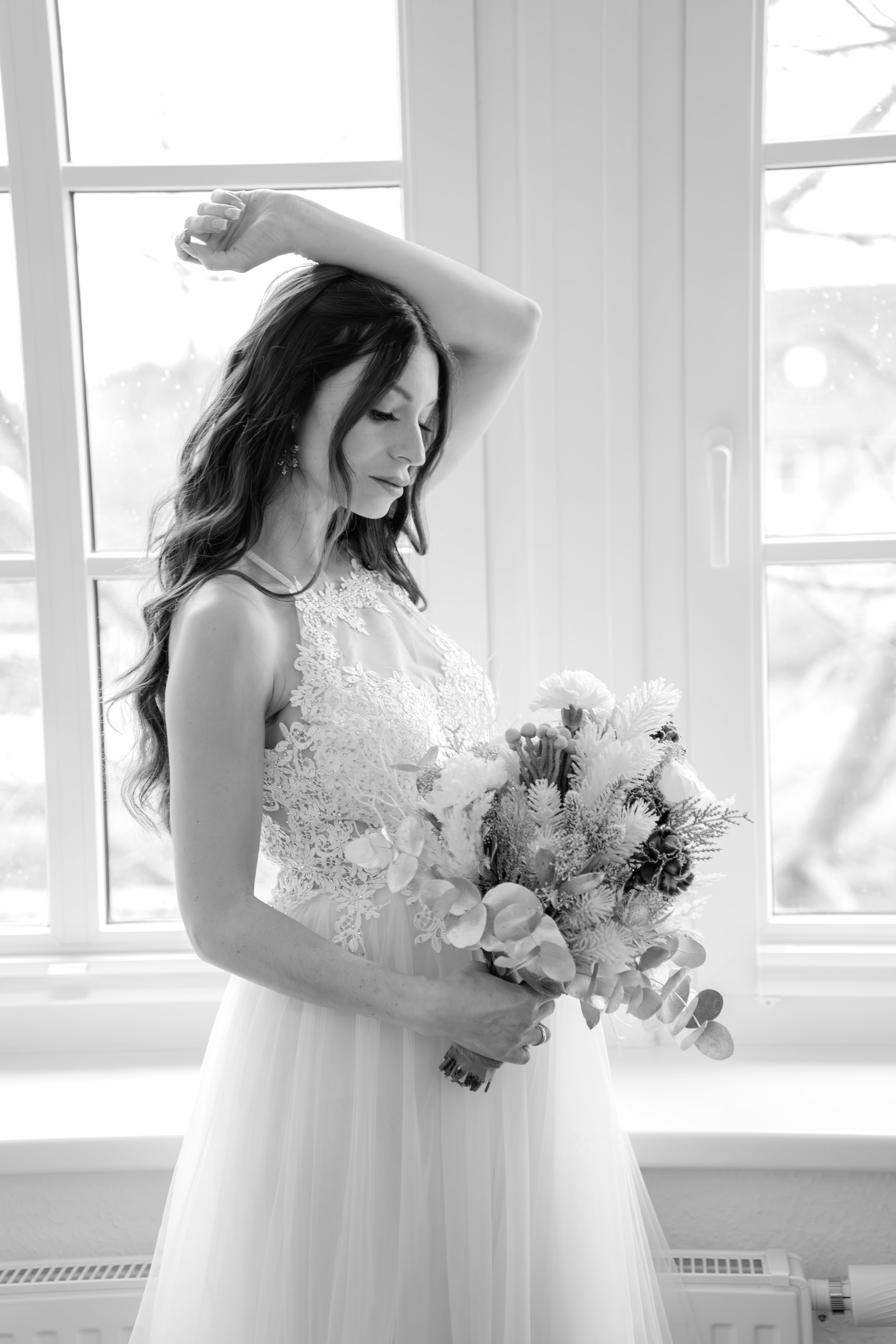 Eine Frau in einem Brautkleid hält einen Blumenstrauß in der Hand vor einem Fenster in einem hell erleuchteten Raum.