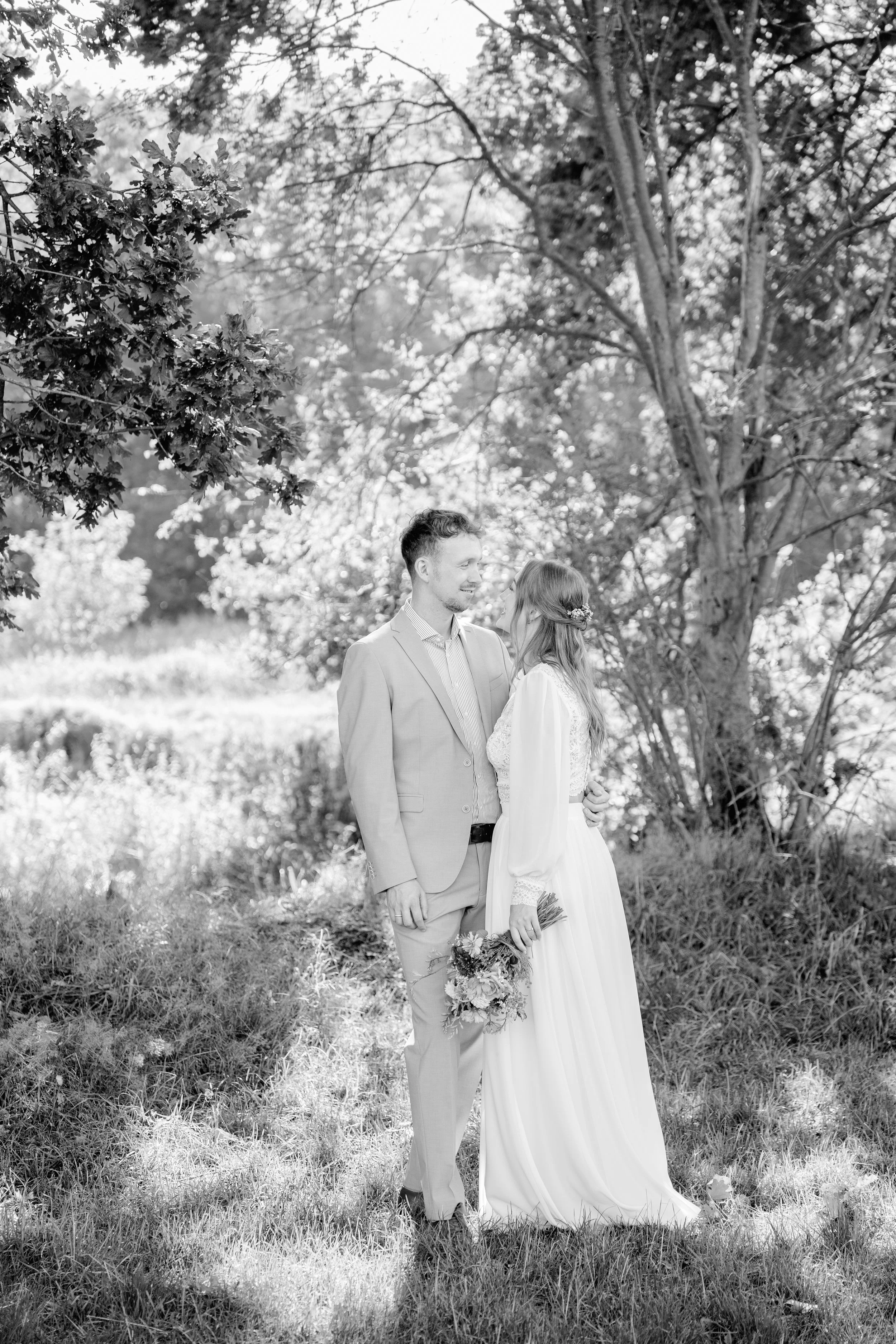 Ein Brautpaar steht sich im Freien gegenüber, umgeben von Bäumen, und lächelt sich liebevoll an. Die Frau hält einen Blumenstrauß in der Hand.
