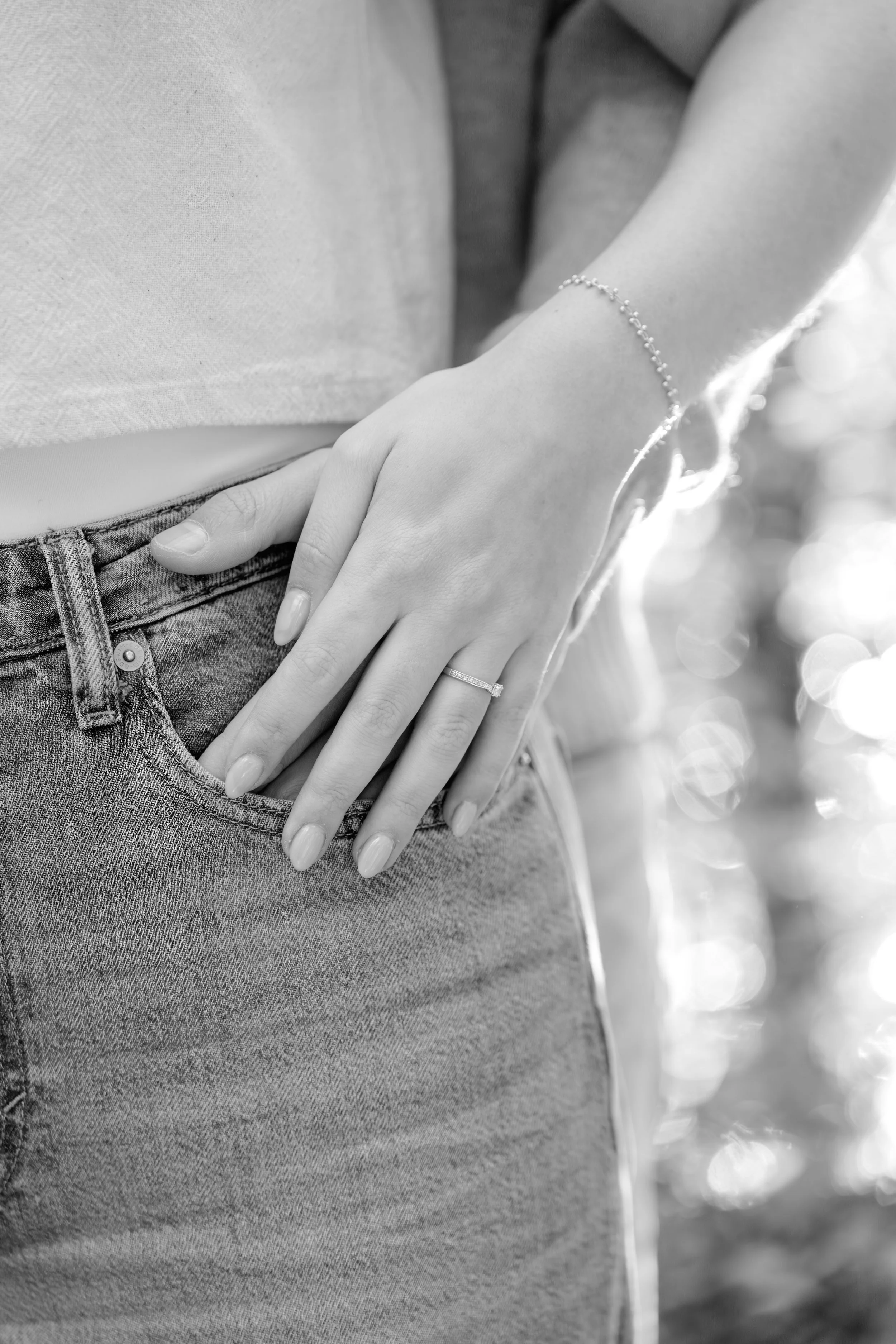 Nahaufnahme einer Hand mit Verlobungsring an der Taille einer Person, in Schwarz-Weiß, mit glänzender Kleidung im Hintergrund.