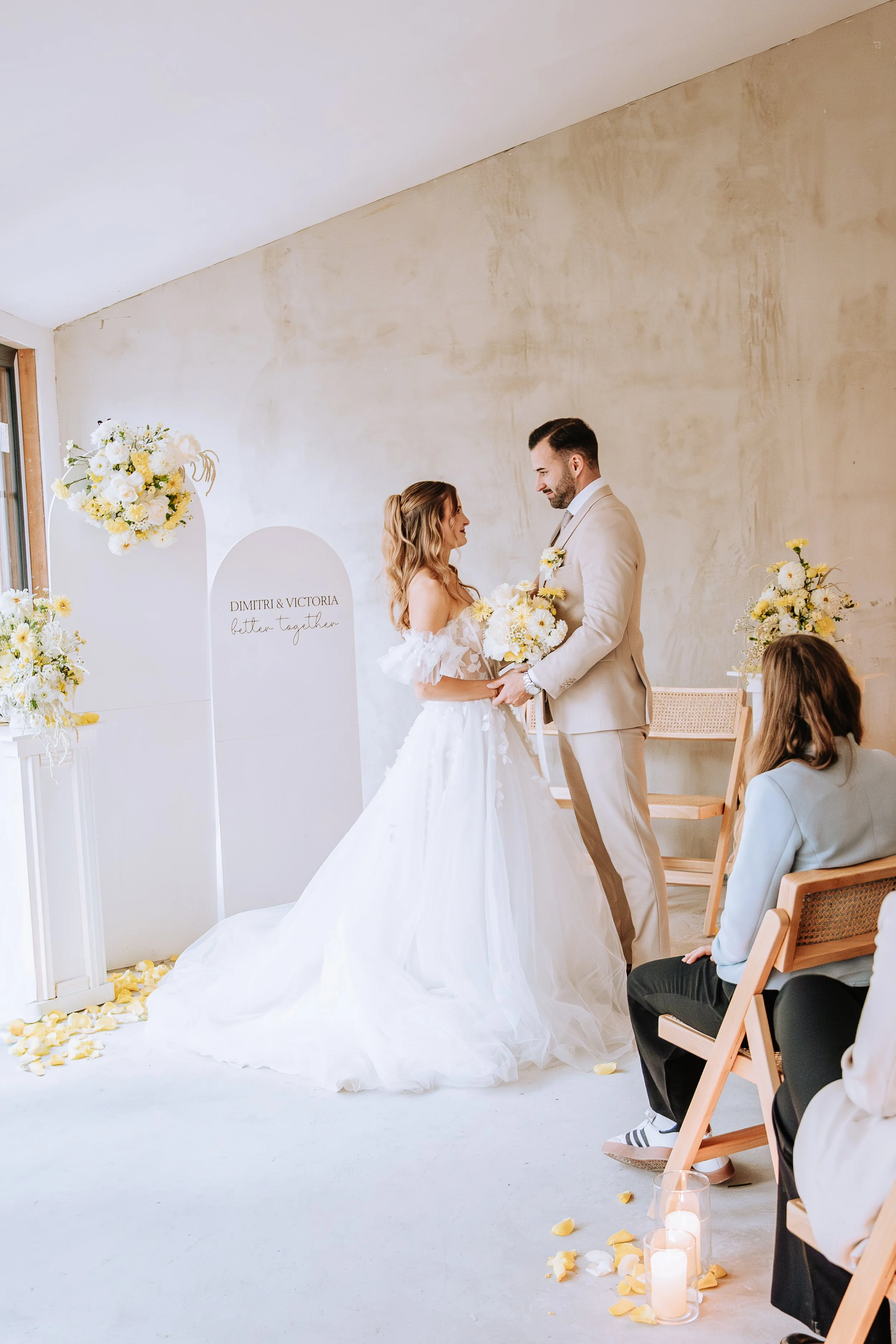 Braut und Bräutigam bei einer Hochzeit stehen sich gegenüber, die Braut trägt ein weißes Hochzeitskleid und der Bräutigam einen hellen Anzug. Es gibt Blumenarrangements und Kerzen im Raum.