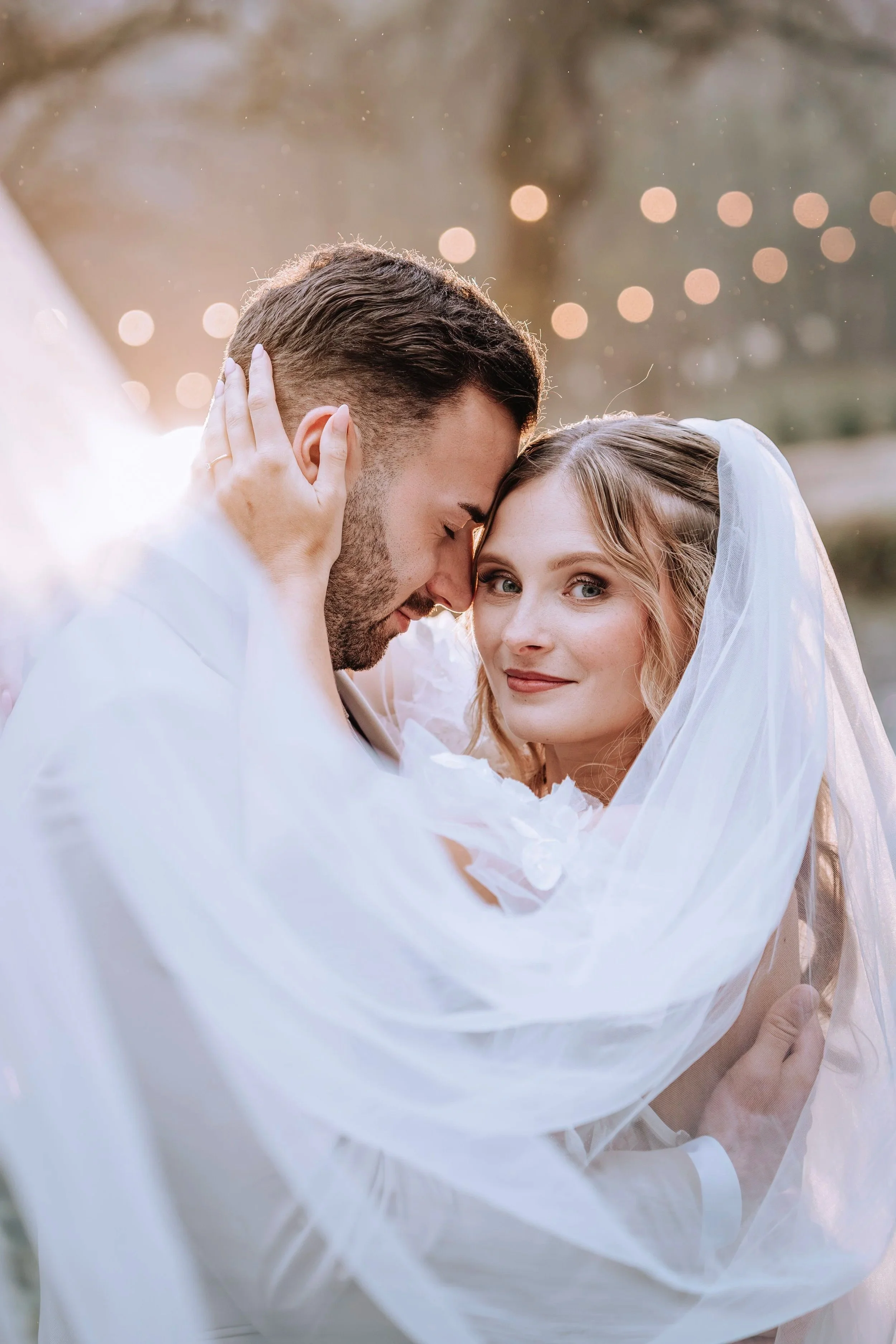 Ein Brautpaar im Hochzeitsoutfit, umschlungen und liebevoll umarmend, bei Sonnenuntergang im Freien, mit unscharfen Lichter im Hintergrund.