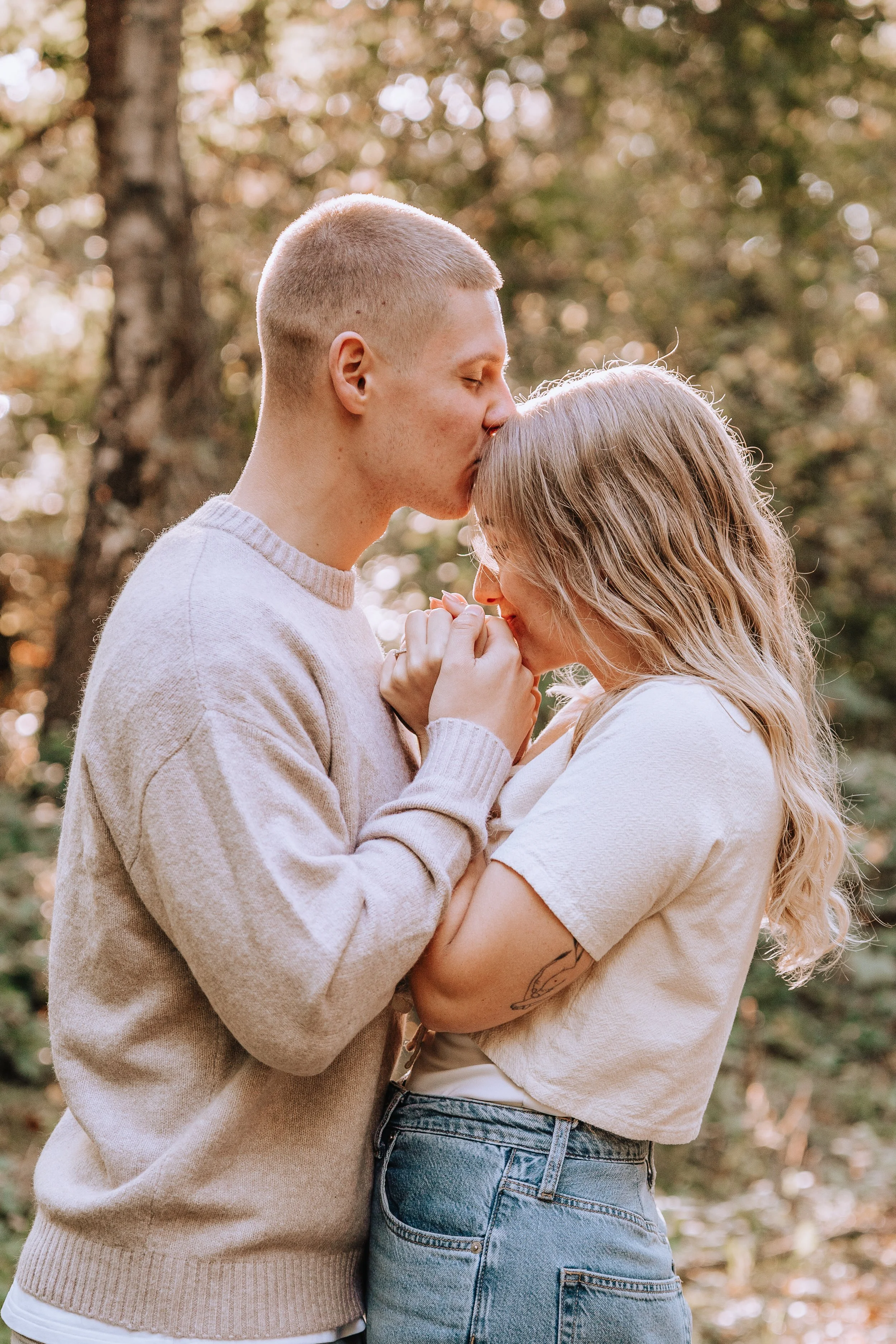 Ein verliebtes Paar umarmt sich im Wald, die Frau hält die Hände des Mannes und sie berühren sich die Stirn. Das Licht scheint durch die Bäume, im Hintergrund sind herbstliche Blätter.