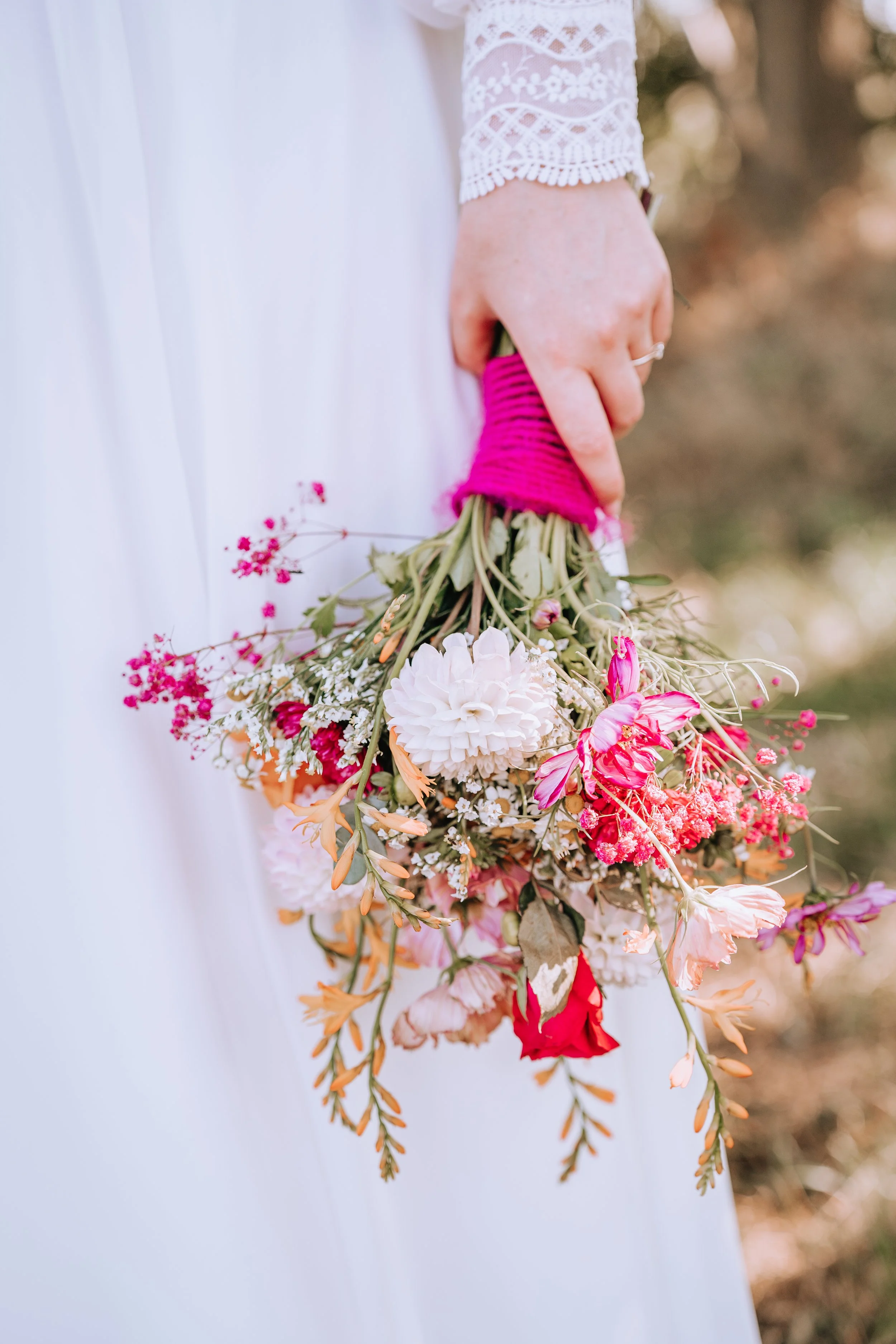 Person in weißem Kleid hält einen bunten Blumenstrauß mit pinkem Wollband um das Bouquet.