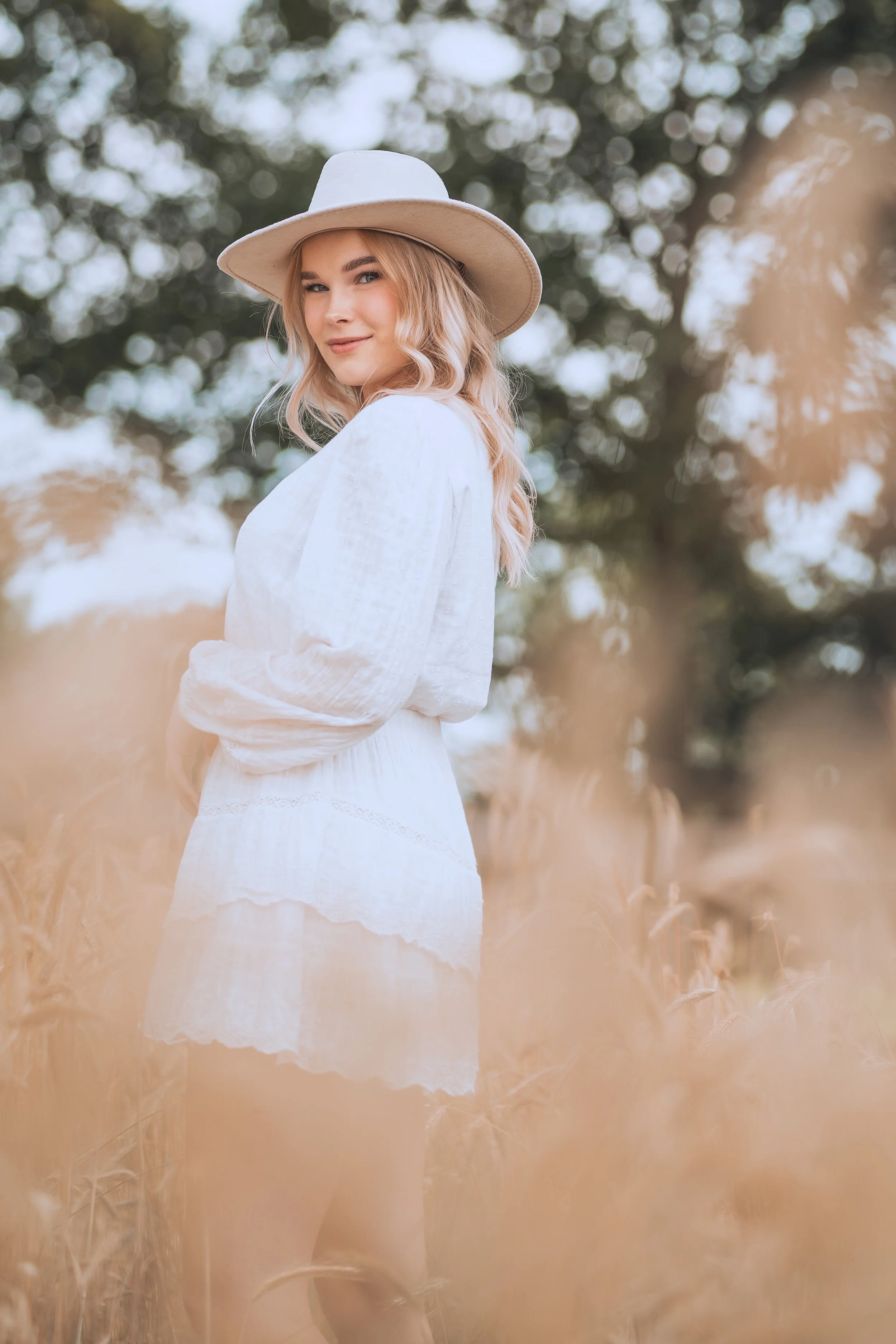 Junge Frau in weißem Kleid und Hut steht in einer Blumenwiese, umgeben von hohen Gräsern, im Sonnenschein, mit einem leichten Lächeln.