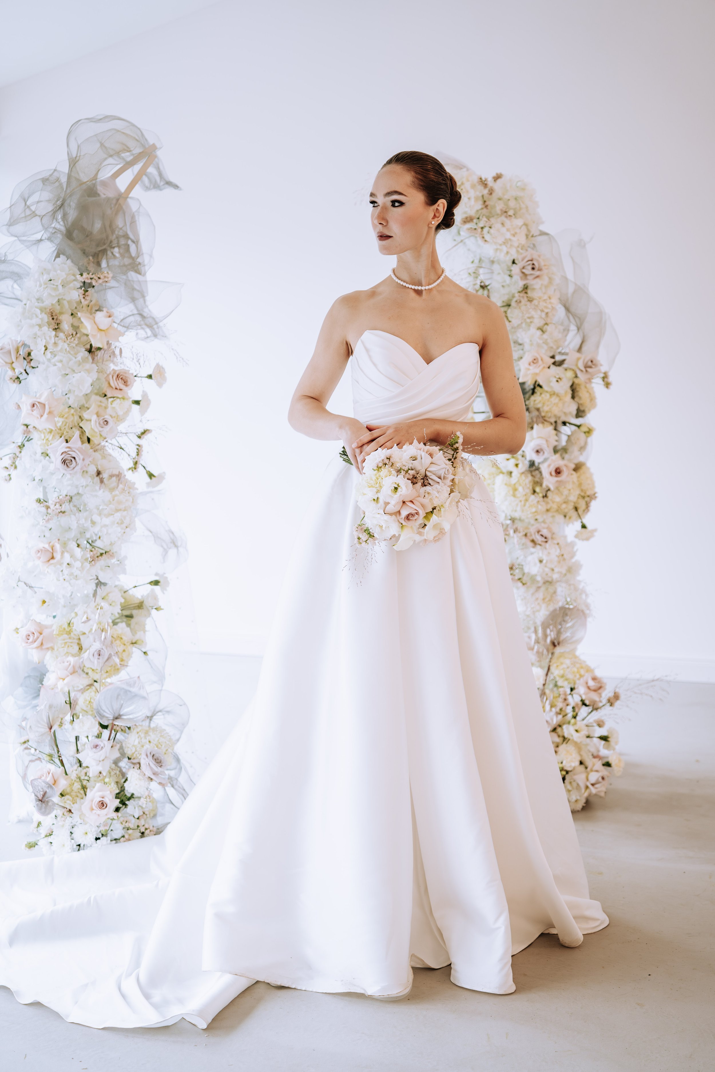 Braut in einem weißen Hochzeitskleid mit Blumenstrauß vor einer Blumendekoration.