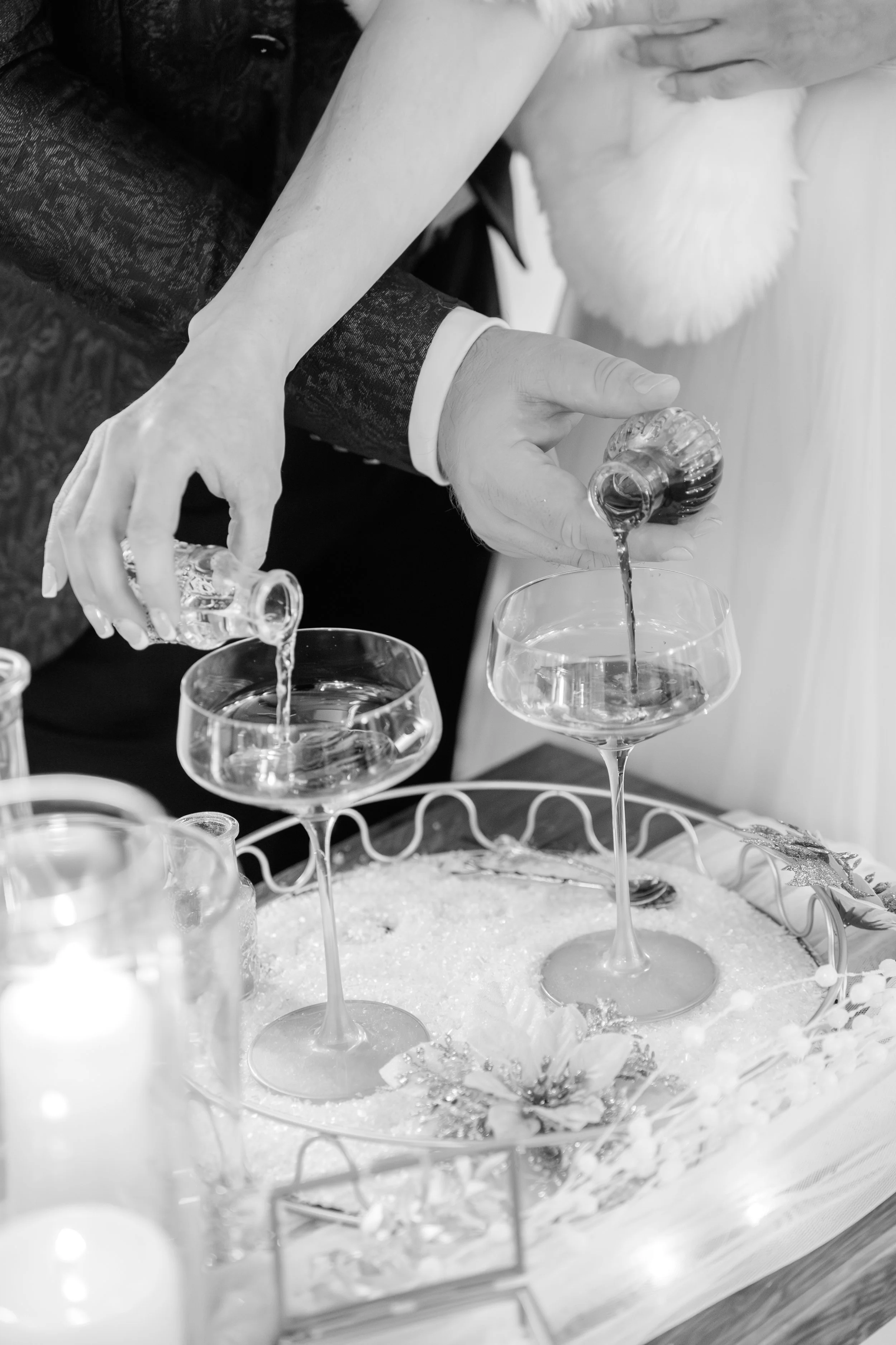 Ein Brautpaar füllt mit Champagner zwei Weingläser bei einer Hochzeitsfeier. Das Bild ist schwarz-weiß.