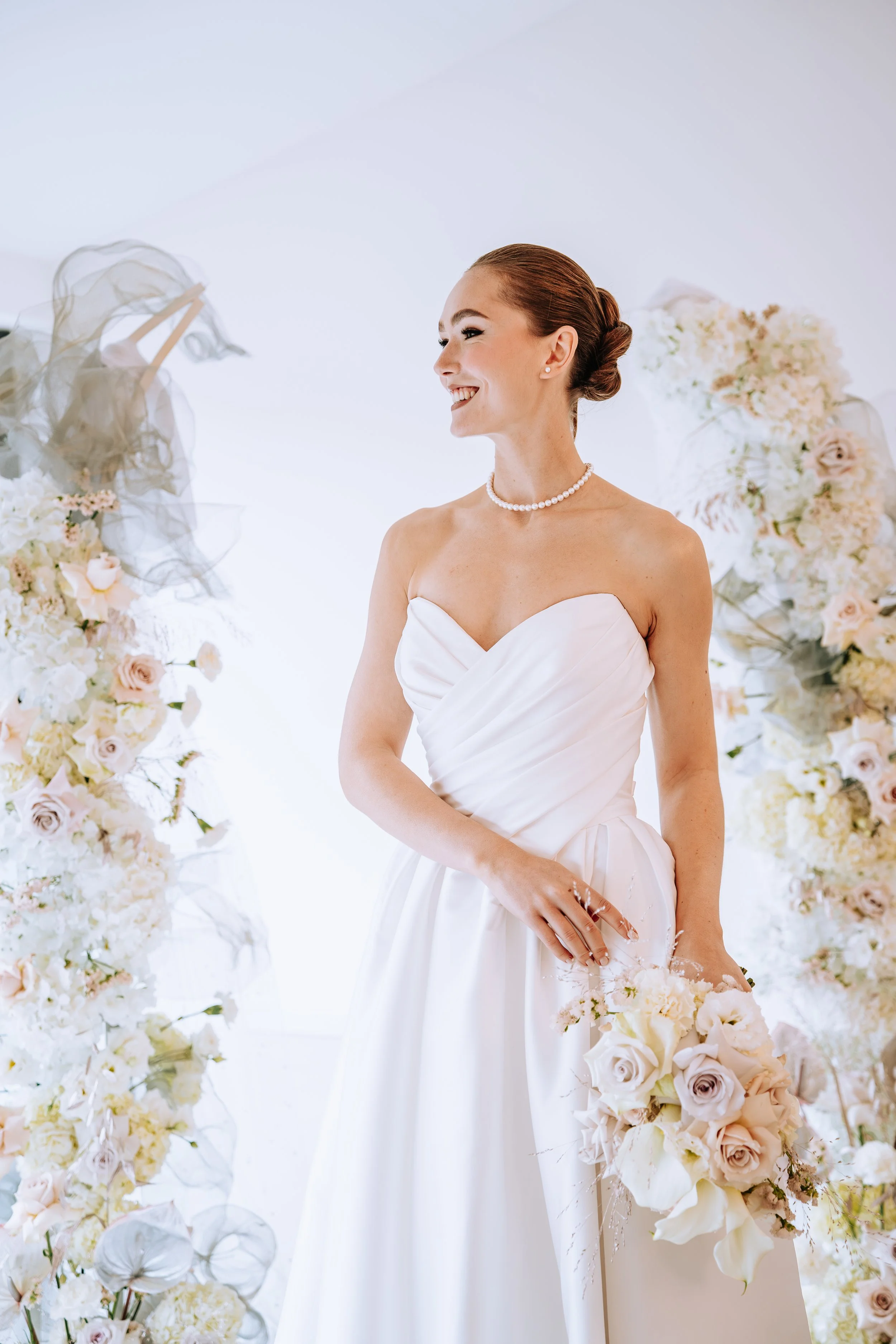 Eine lächelnde Braut in einem weißen Brautkleid, die einen Blumenstrauß hält, umgeben von floraler Dekoration.