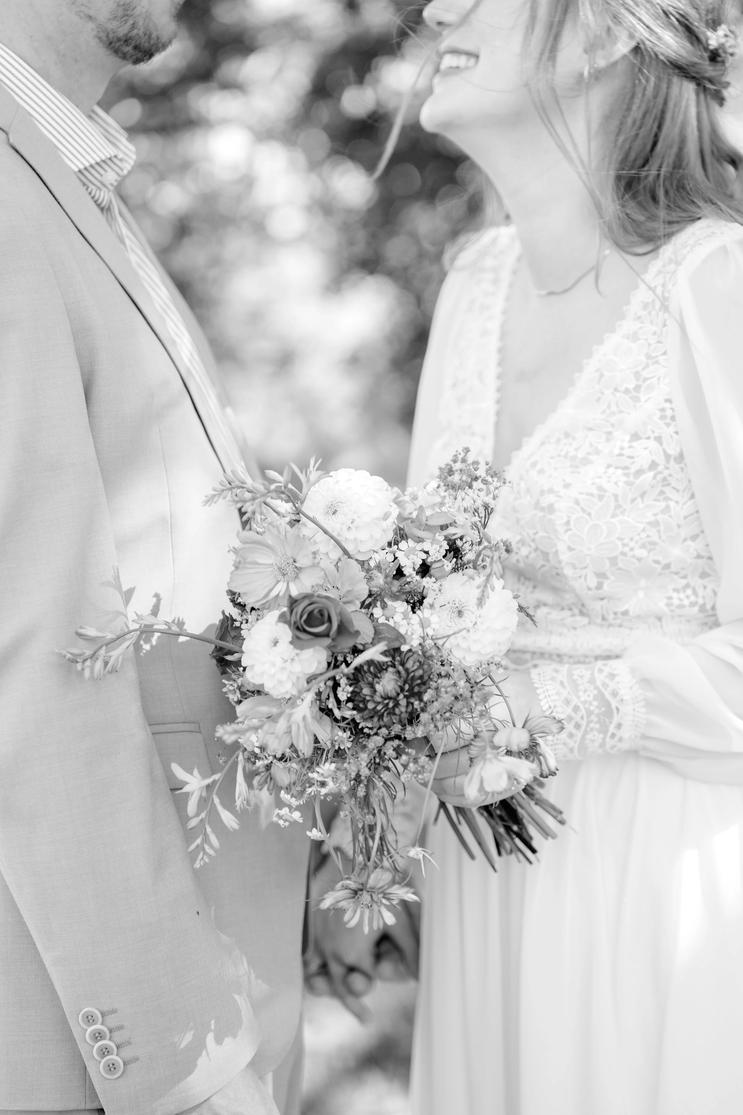 Ein Brautpaar bei einer Hochzeit, die Frau hält einen Blumenstrauß, sie tragen formelle Kleidung, im Hintergrund unscharfe Bäume.