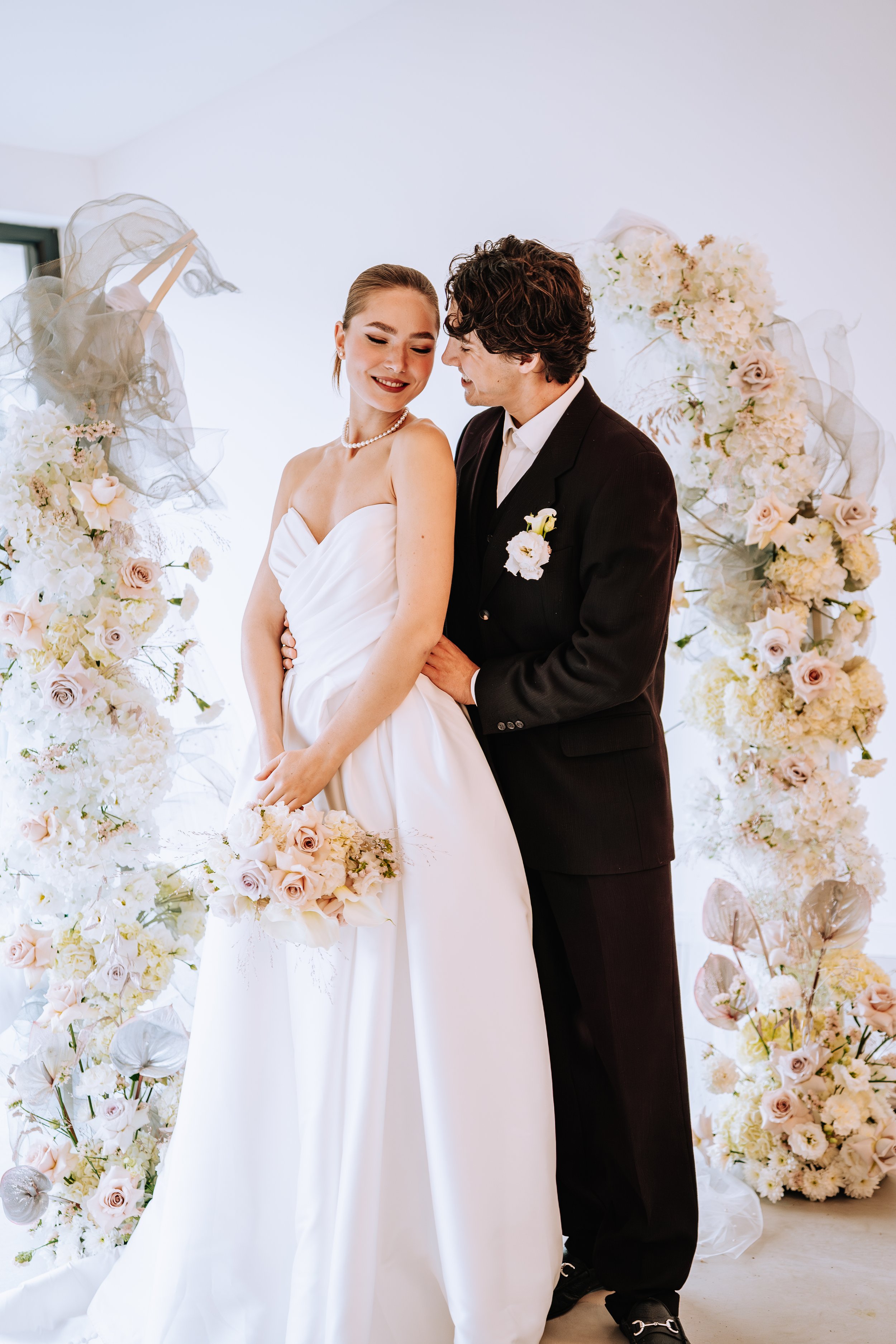 Ein Brautpaar bei ihrer Hochzeit, die Braut trägt ein weißes Kleid und hält einen Brautstrauß, der Bräutigam trägt einen dunklen Anzug, im Hintergrund sind Blumen und Dekorationen.