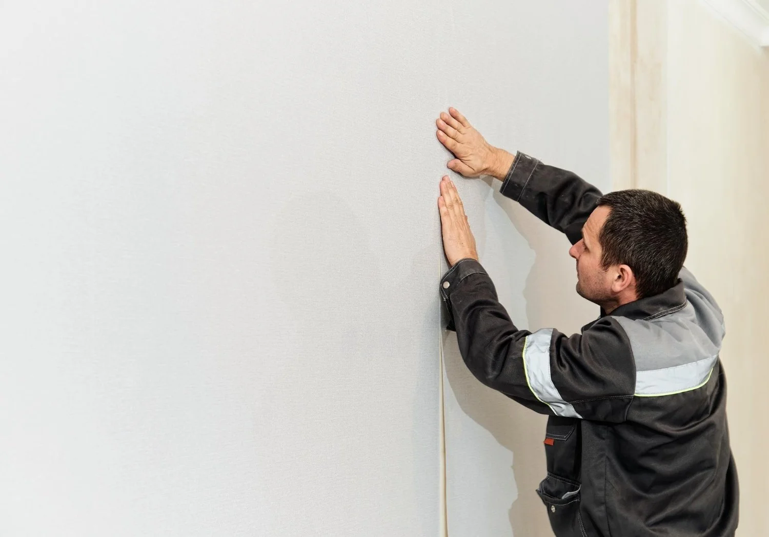 A man in a work jacket is applying wallpaper to a wall in a room.