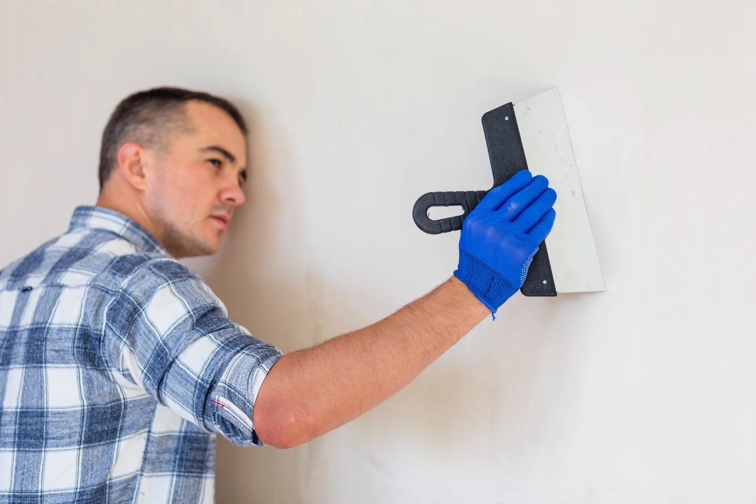 A man in a plaid shirt and blue gloves using a trowel to apply joint compound or spackle to a wall.