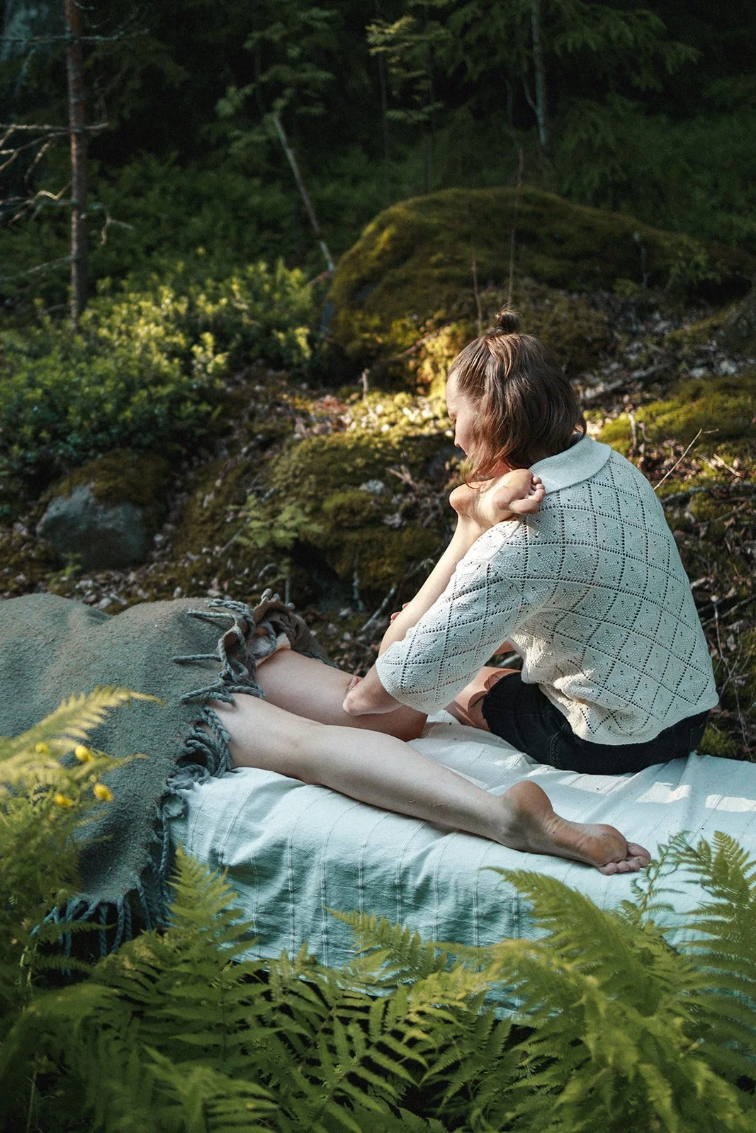 A woman is sitting on a bed or platform in a forest, wearing a white crochet top and black shorts, with her knees drawn up, gently holding and looking at her feet, surrounded by green foliage and moss-covered rocks.