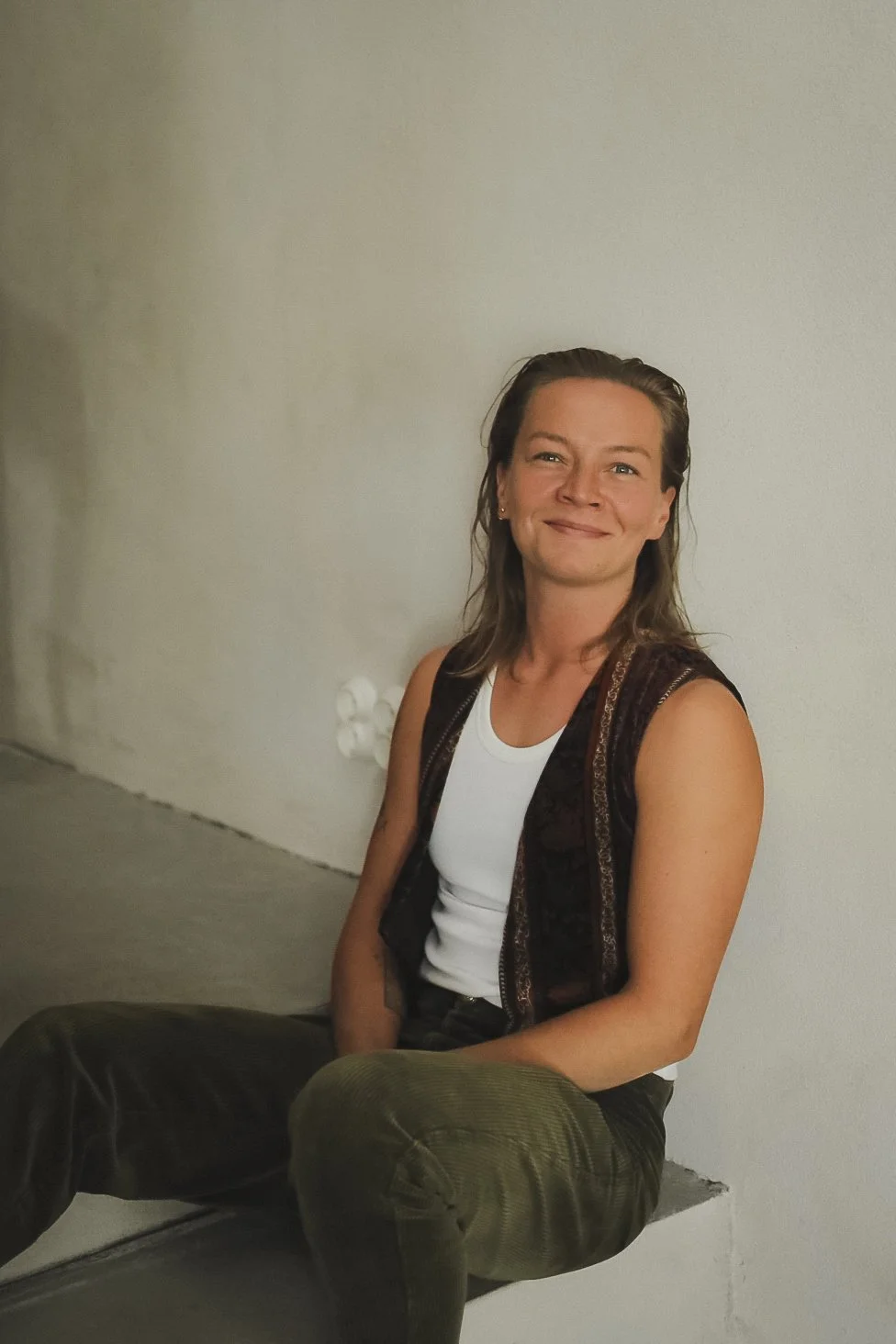 A woman with shoulder-length brown hair sitting against a plain white wall, smiling, wearing a white tank top, a dark patterned vest, and green pants.