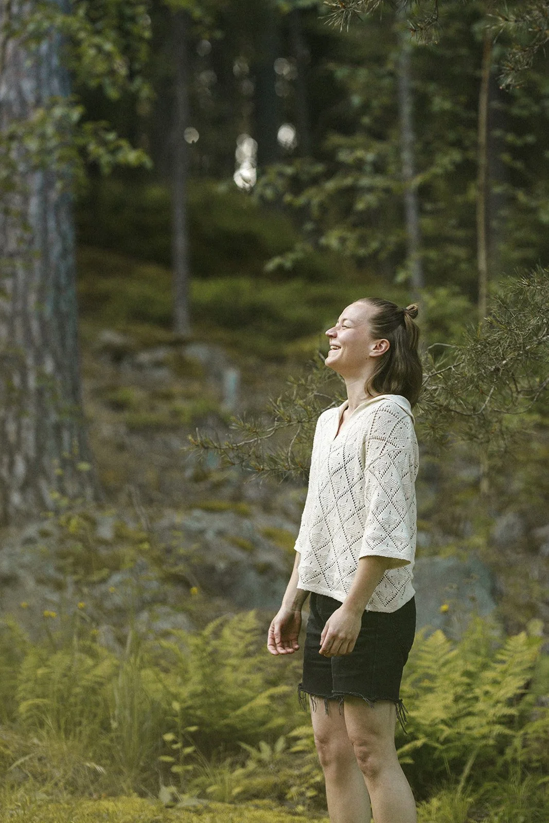 A woman standing outdoors in a forest, smiling and laughing, wearing a cream-colored knit sweater and black shorts, surrounded by green trees and foliage.