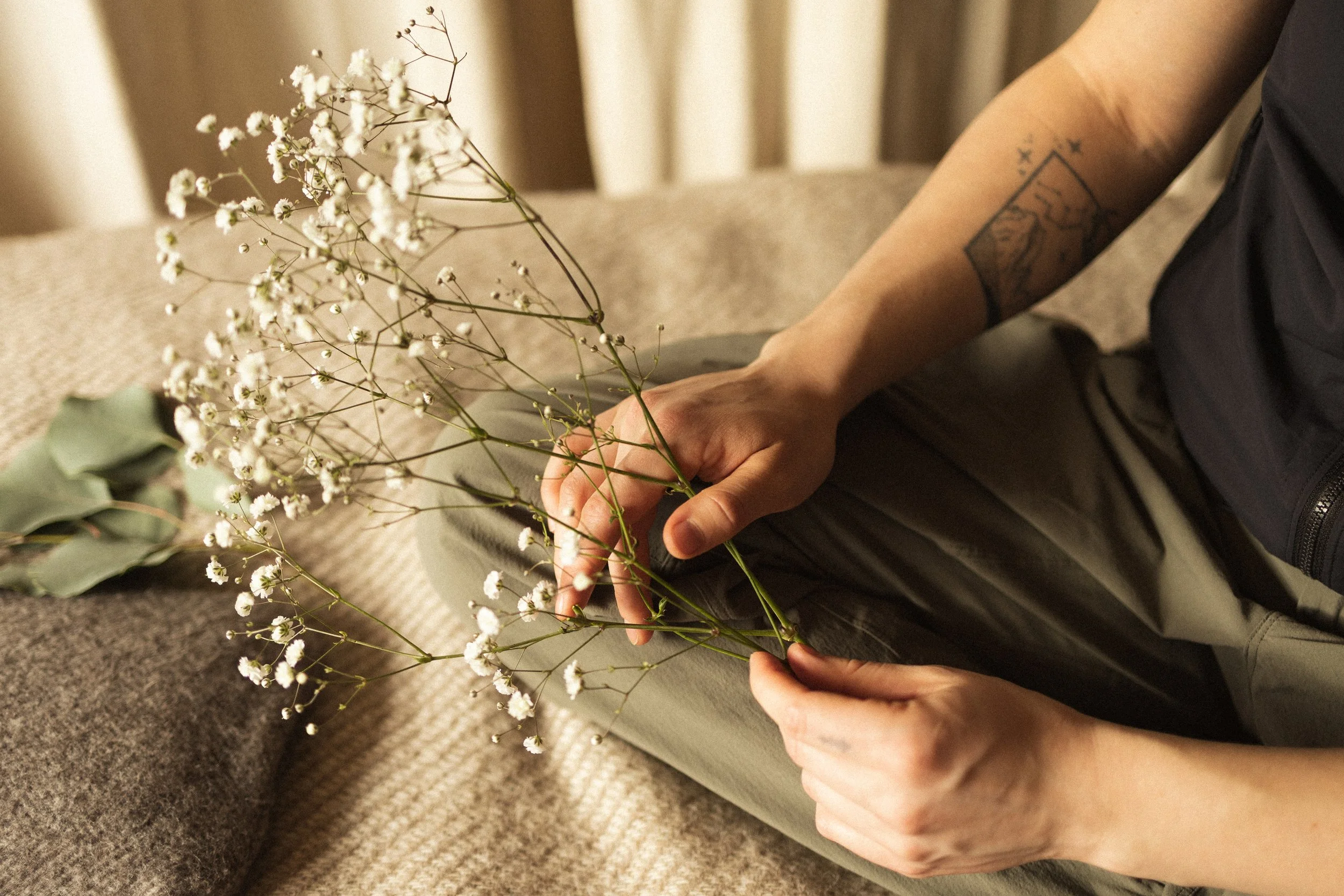 Person sitting on carpeted floor holding small white flowers with green stems and leaves, tattoo on arm, wearing gray pants and black jacket.