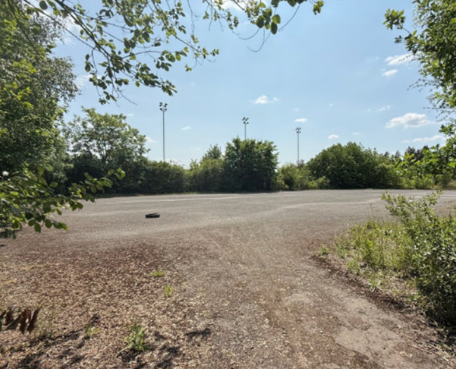 Empty parking lot with a tire in the middle, surrounded by trees and bushes, with tall light poles under a partly cloudy sky.
