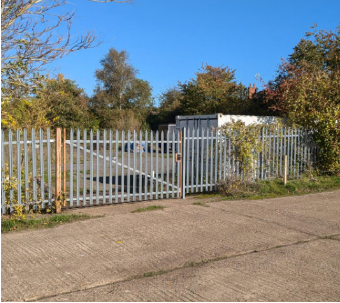 A metal gate and fence along a sidewalk, with trees and a clear blue sky in the background.