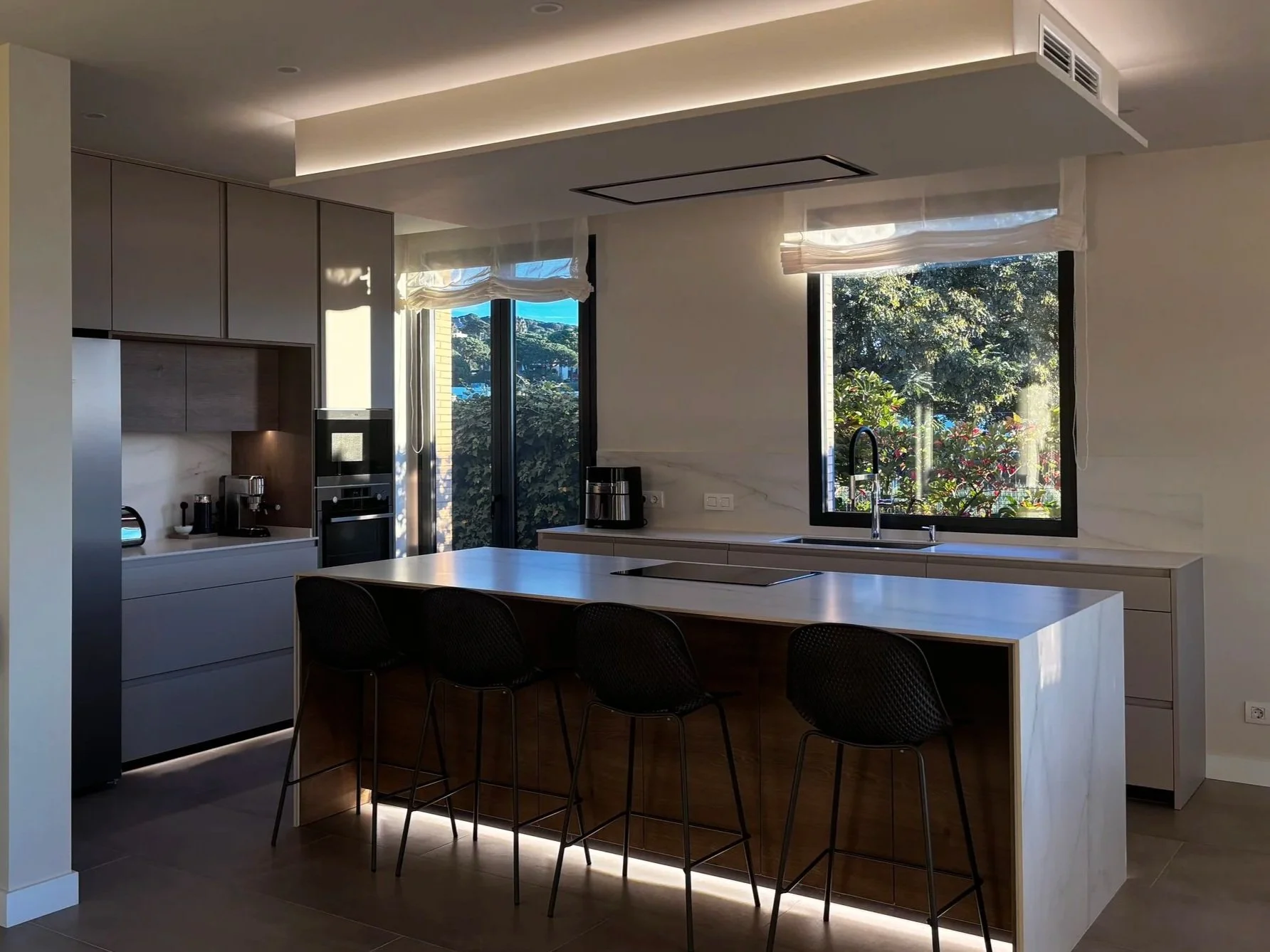 Cocina moderna con encimera de isla de madera, despensa gris, ventanas grandes con vista a un jardín y sillas negras en la isla.