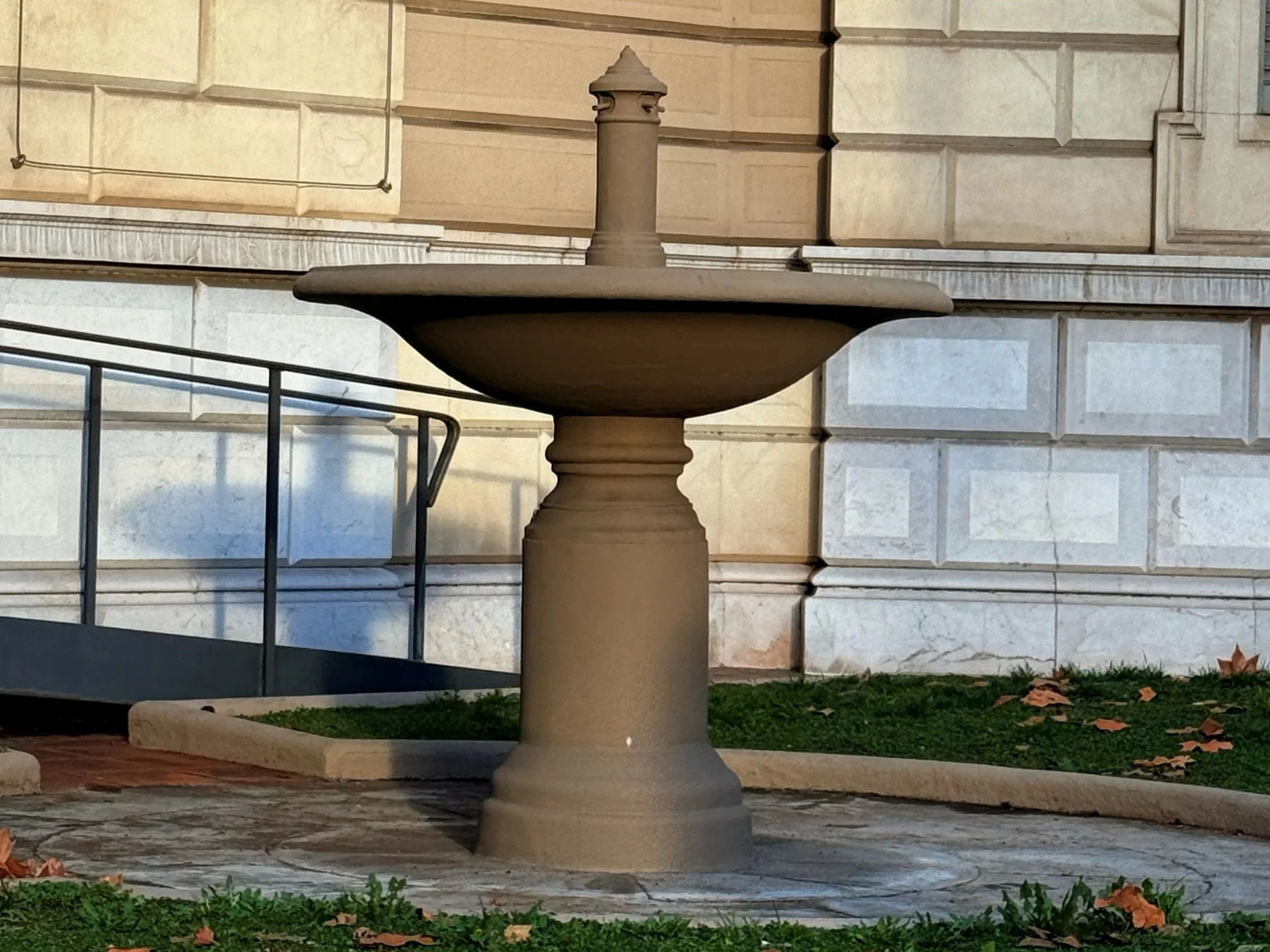 Fuente de piedra en un área exterior, con un edificio de piedra en el fondo, con hojas caídas en el suelo cercano.