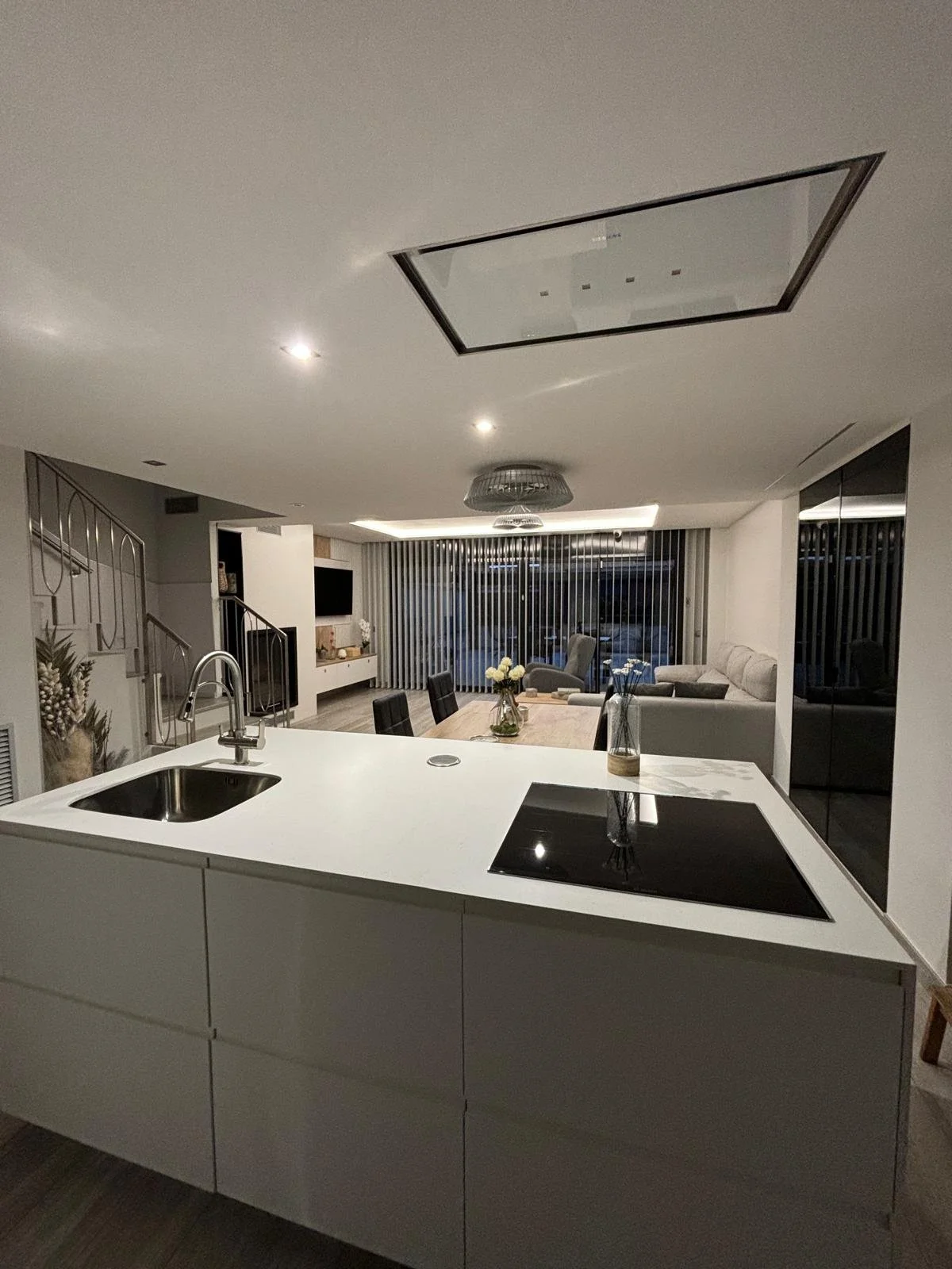 Vista de una cocina moderna con isla blanca, estufa eléctrica y fregadero, vista hacia la sala con sillones y mesas con flores, en un ambiente de iluminación tenue.
