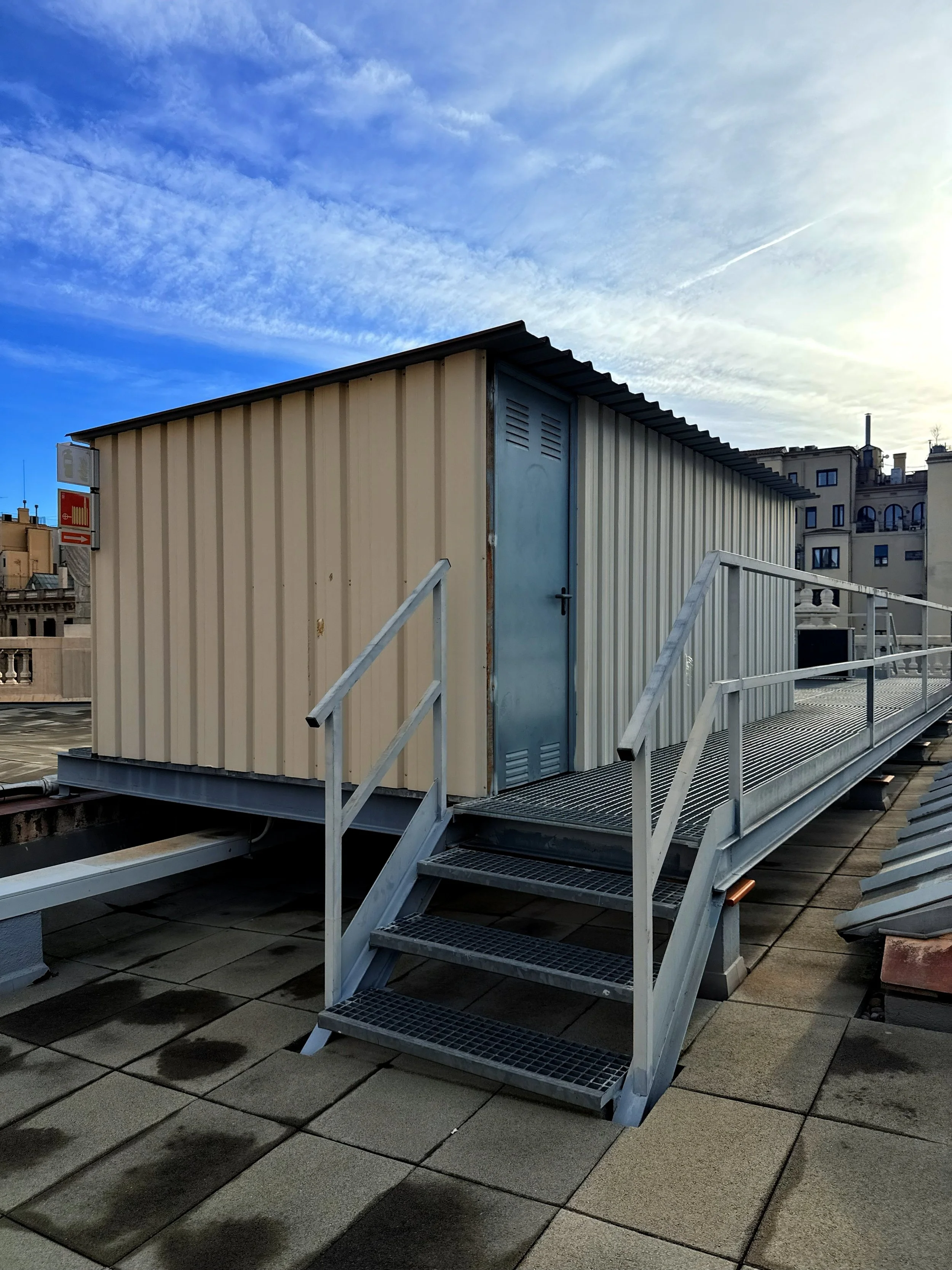 Edificio pequeño metálico en la azotea con escaleras y rampa de acceso, con vista de edificios y cielo azul con nubes.