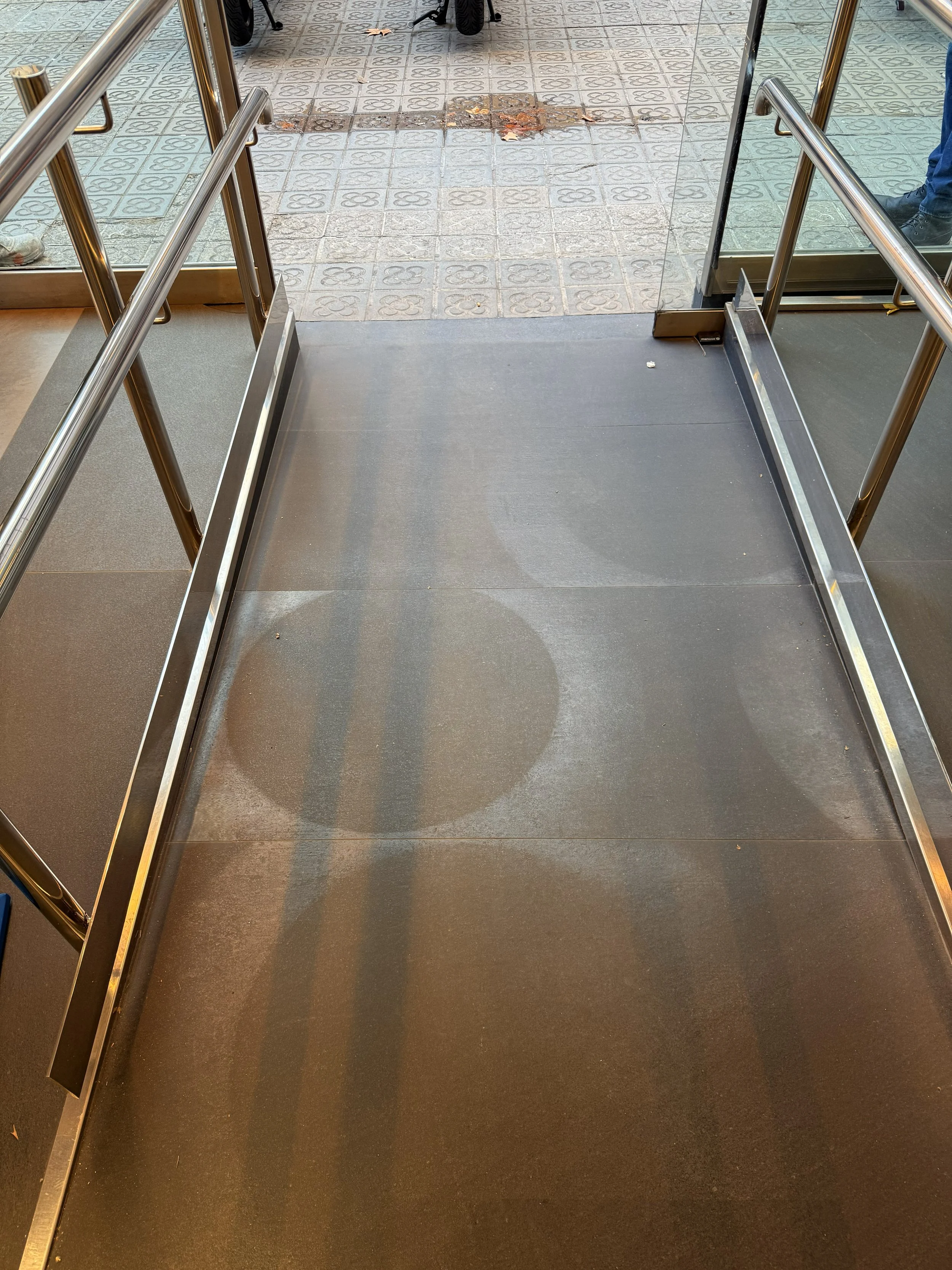 Entrada de un local con piso de metal y pasamanos de acero, vista del umbral con suelo de baldosas y hojas caídas afuera.