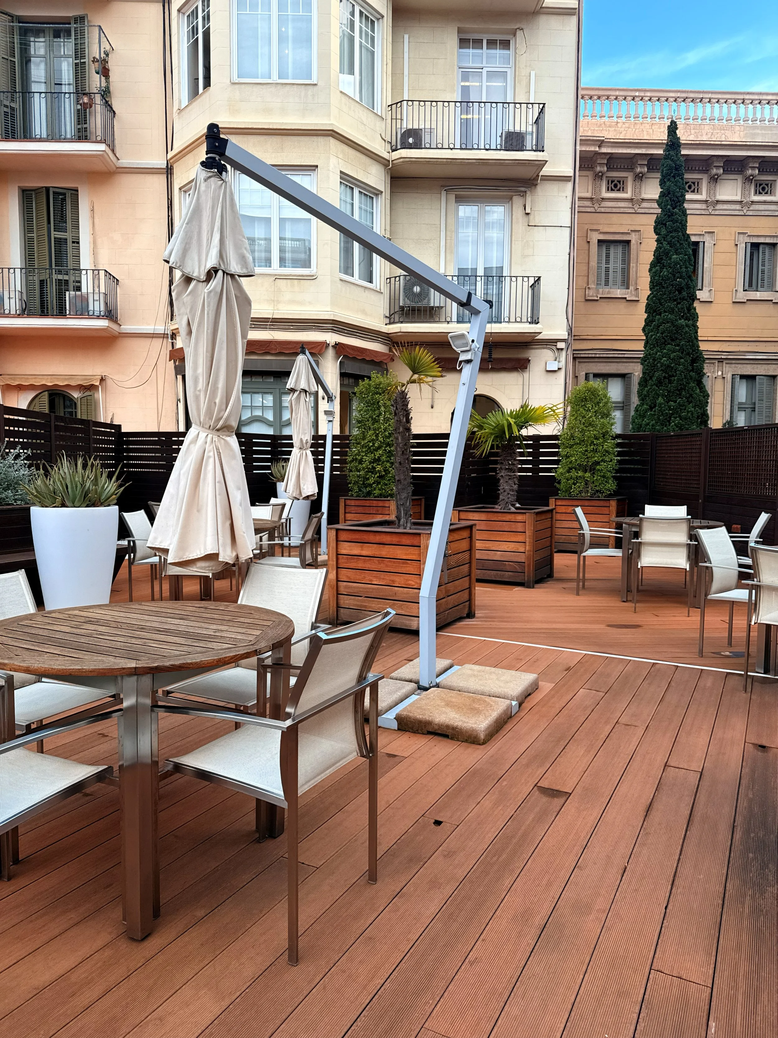 Terraza con mesas y sillas, toldo plegado, plantas en jardineras de madera, edificios residenciales en fondo, cielo despejado.
