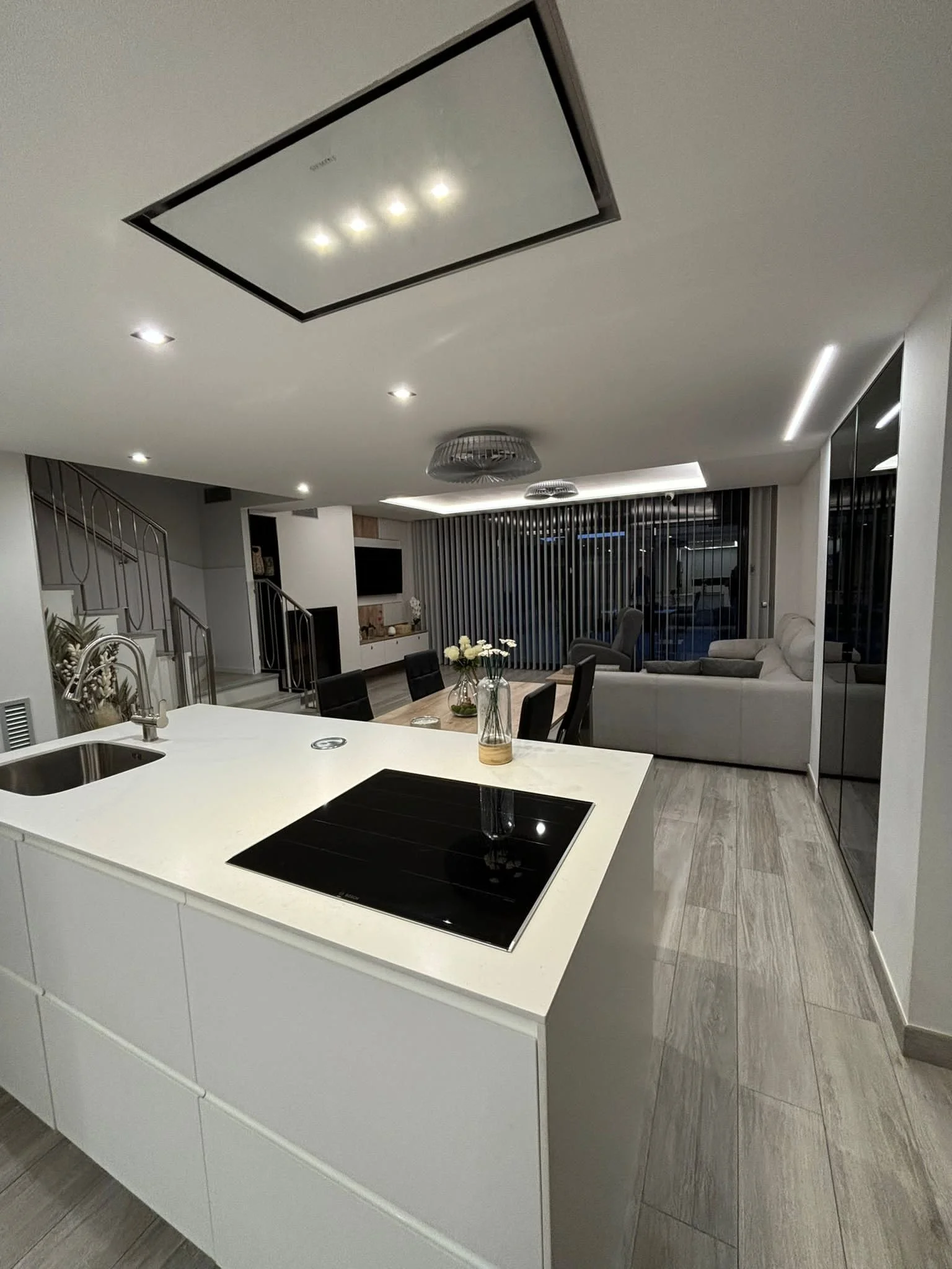 Vista de una casa moderna con cocina, comedor y sala, iluminada con luces empotradas y decorada con flores.