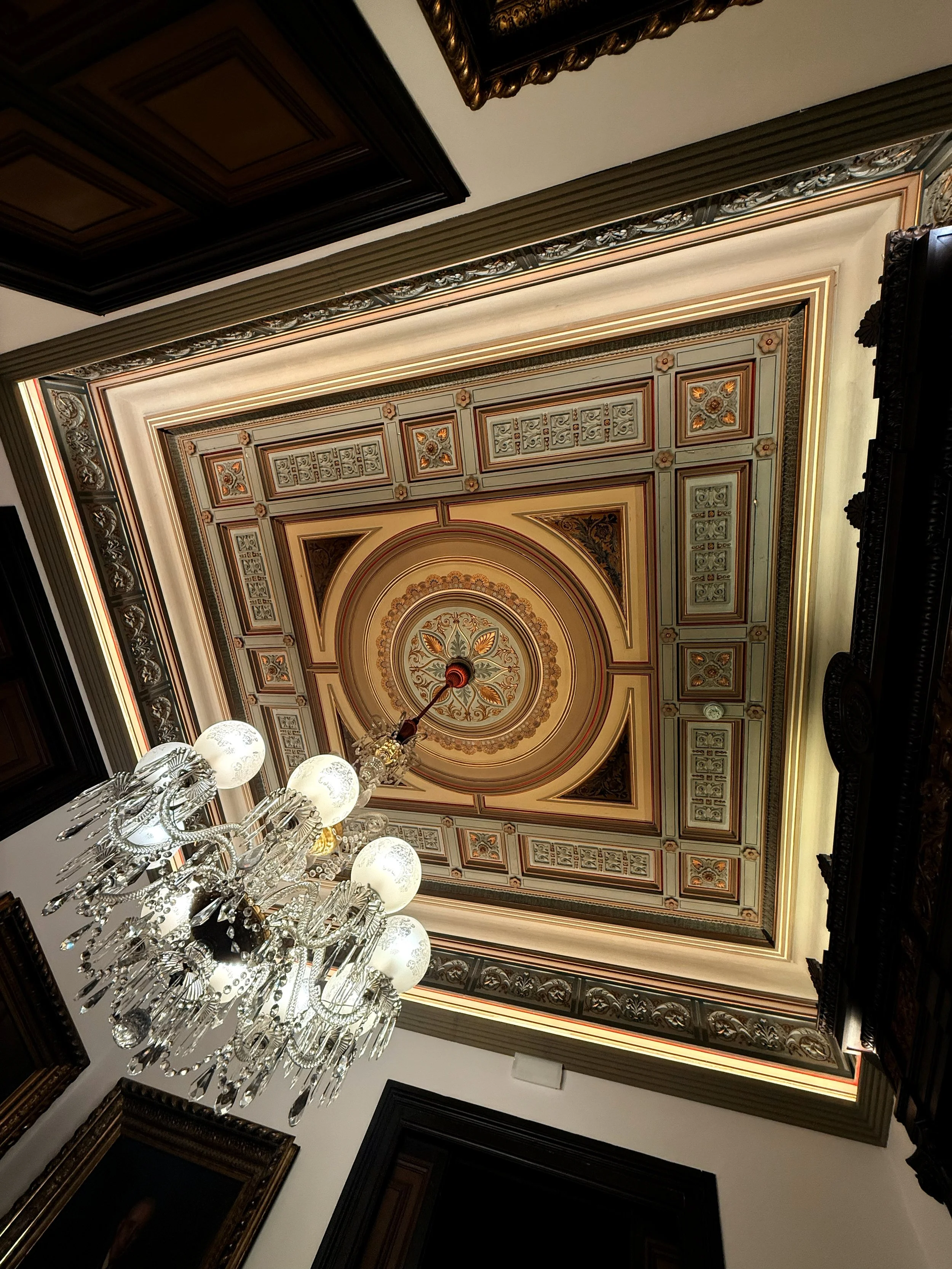 Techo decorado con molduras ornamentales, en el centro un medallón con diseño floral, y un candelabro de cristales con varias globos de luz colgado del centro. 