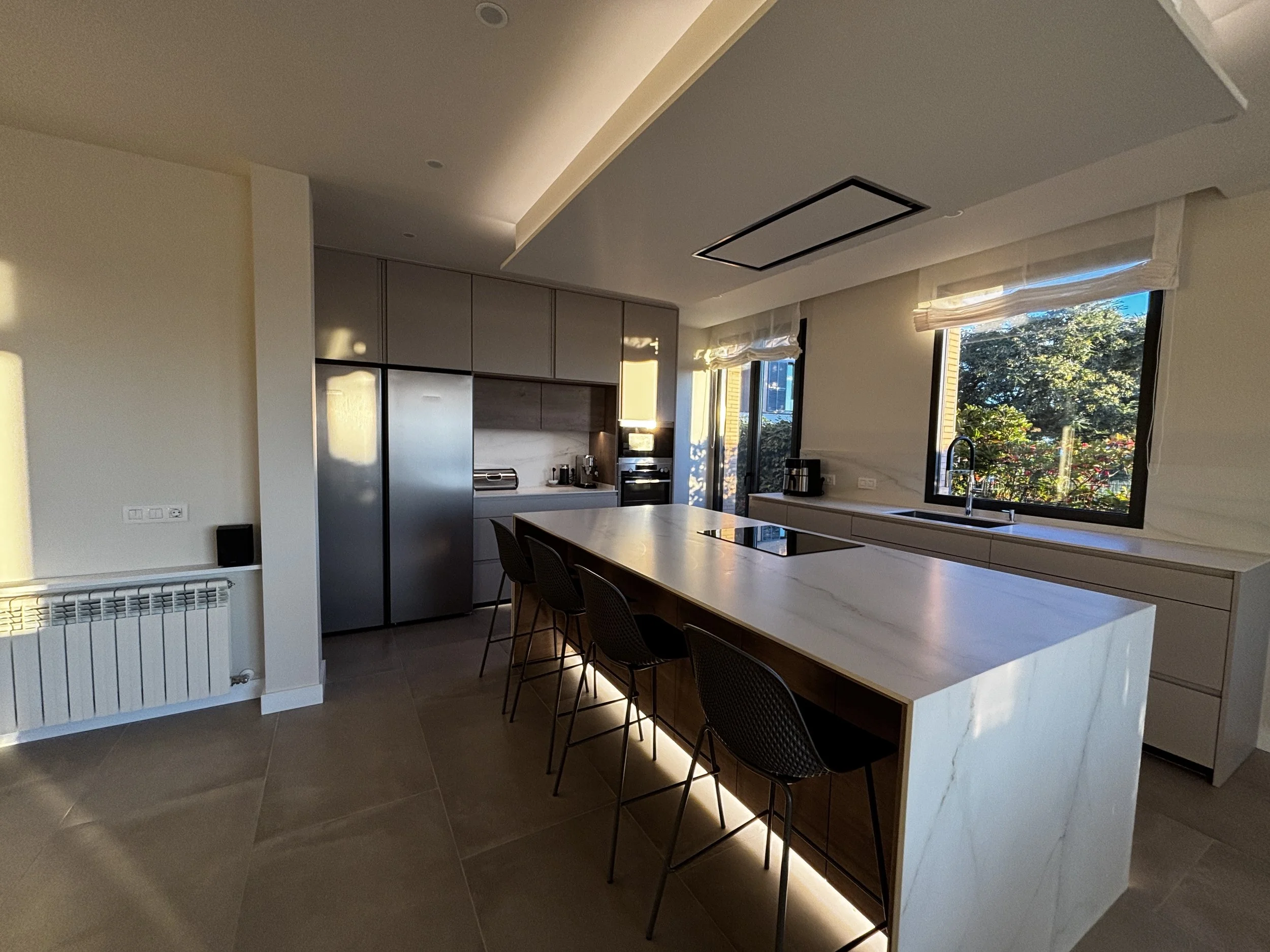 Cocina moderna con isla de mármol, ventana grande con vista a un jardín, electrodomésticos de acero inoxidable, y barra con sillas negras.