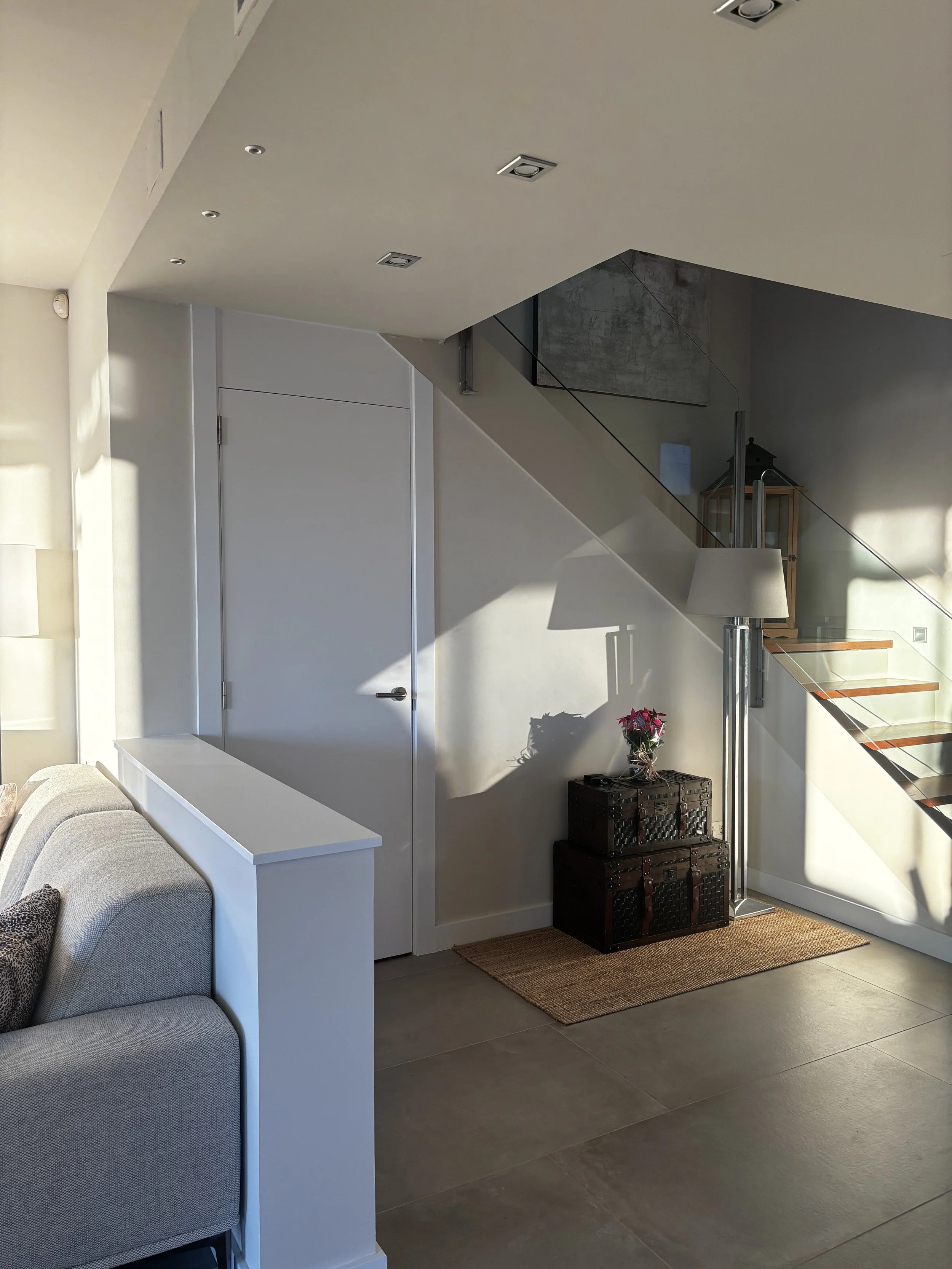 Salón moderno con una escalera de cristal y madera, una lámpara de pie junto a dos maletas negras con una maceta con flores rosadas encima, y un tapete de yute en el piso.