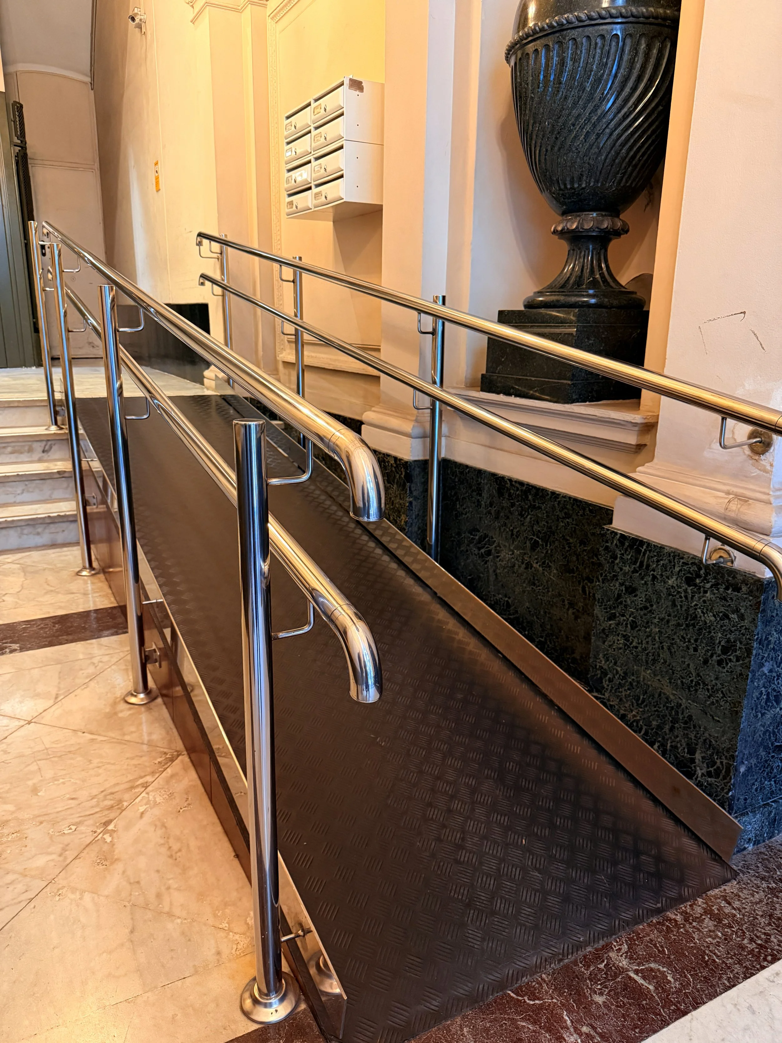 Rampa de acceso para discapacitados en el interior de un edificio, con barandales de metal y suelo de goma negra.
