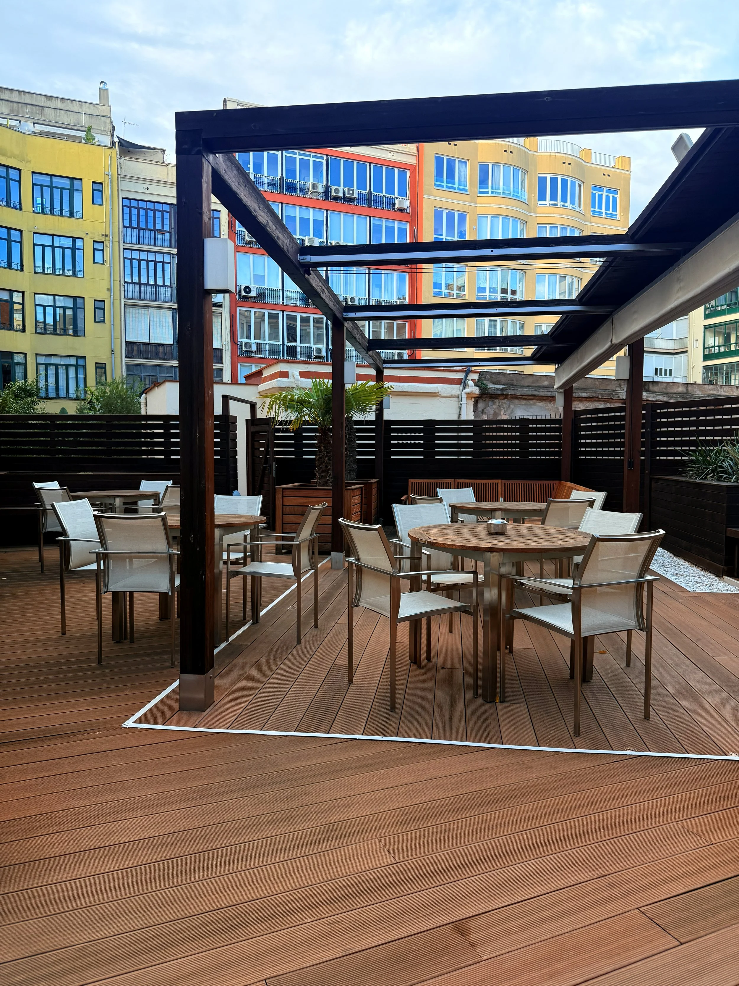 Terraza de restaurante con mesas de madera, sillas de metal y un área techada sin techo en un entorno urbano.