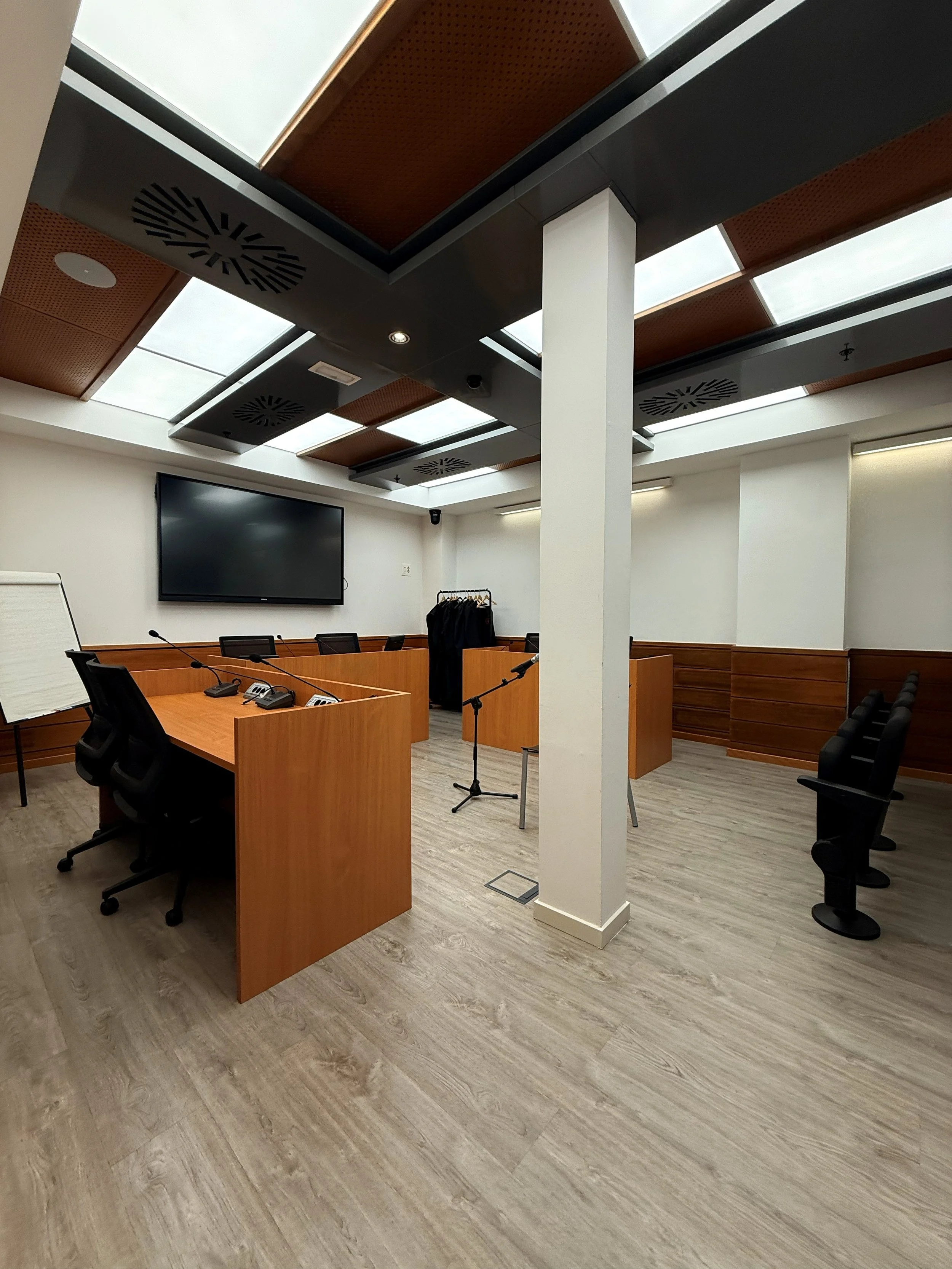 Sala de reuniones moderna y vacía con mesas de madera, sillas ergonómicas, pantalla grande en la pared, micrófonos, una pizarra de caballete y un vestidor con ropa colgada.