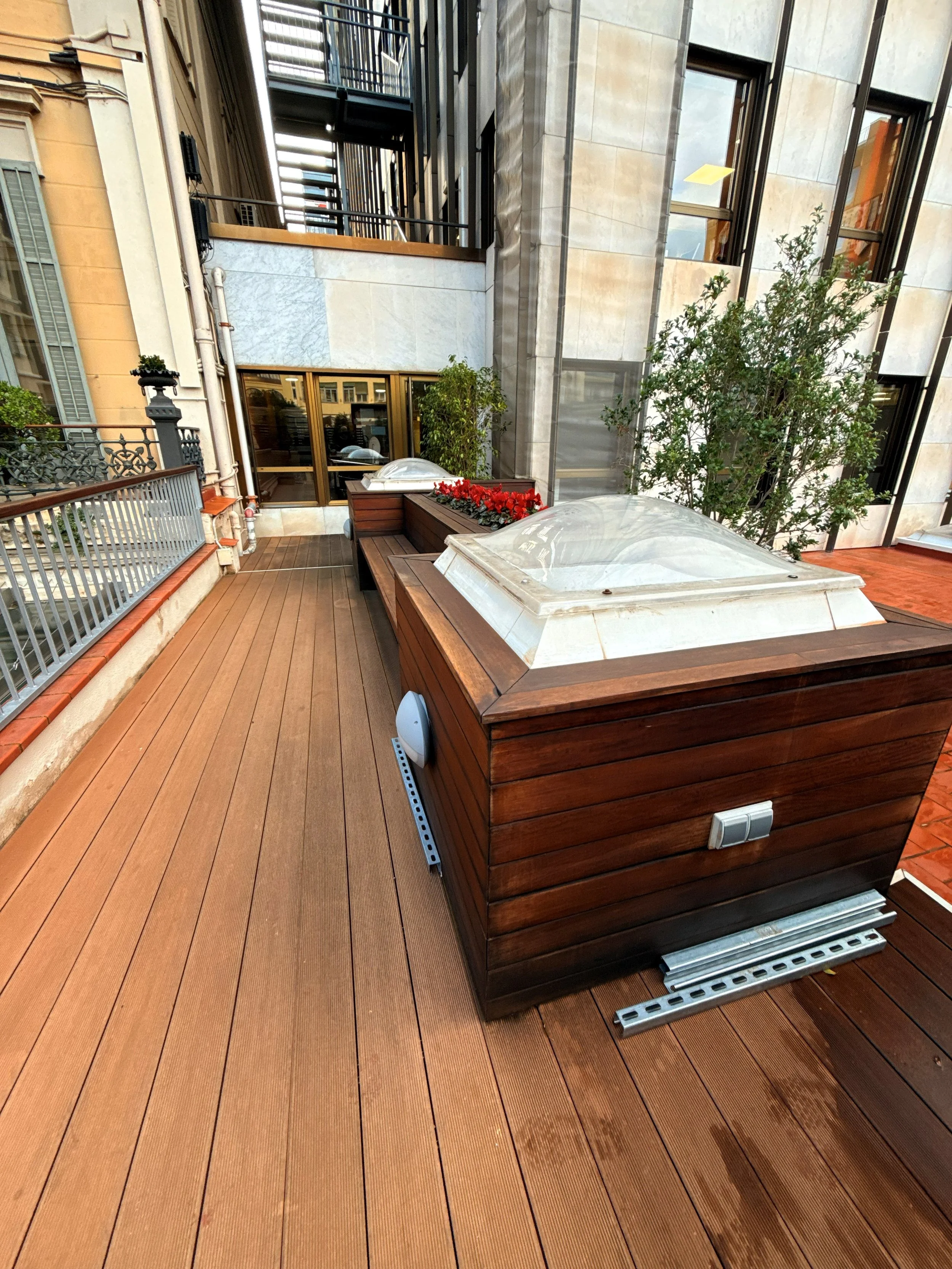 Terraza de edificio con madera en el suelo, dos lucernarios en remates de madera, plantas y ventanas de vidrio en la fachada.
